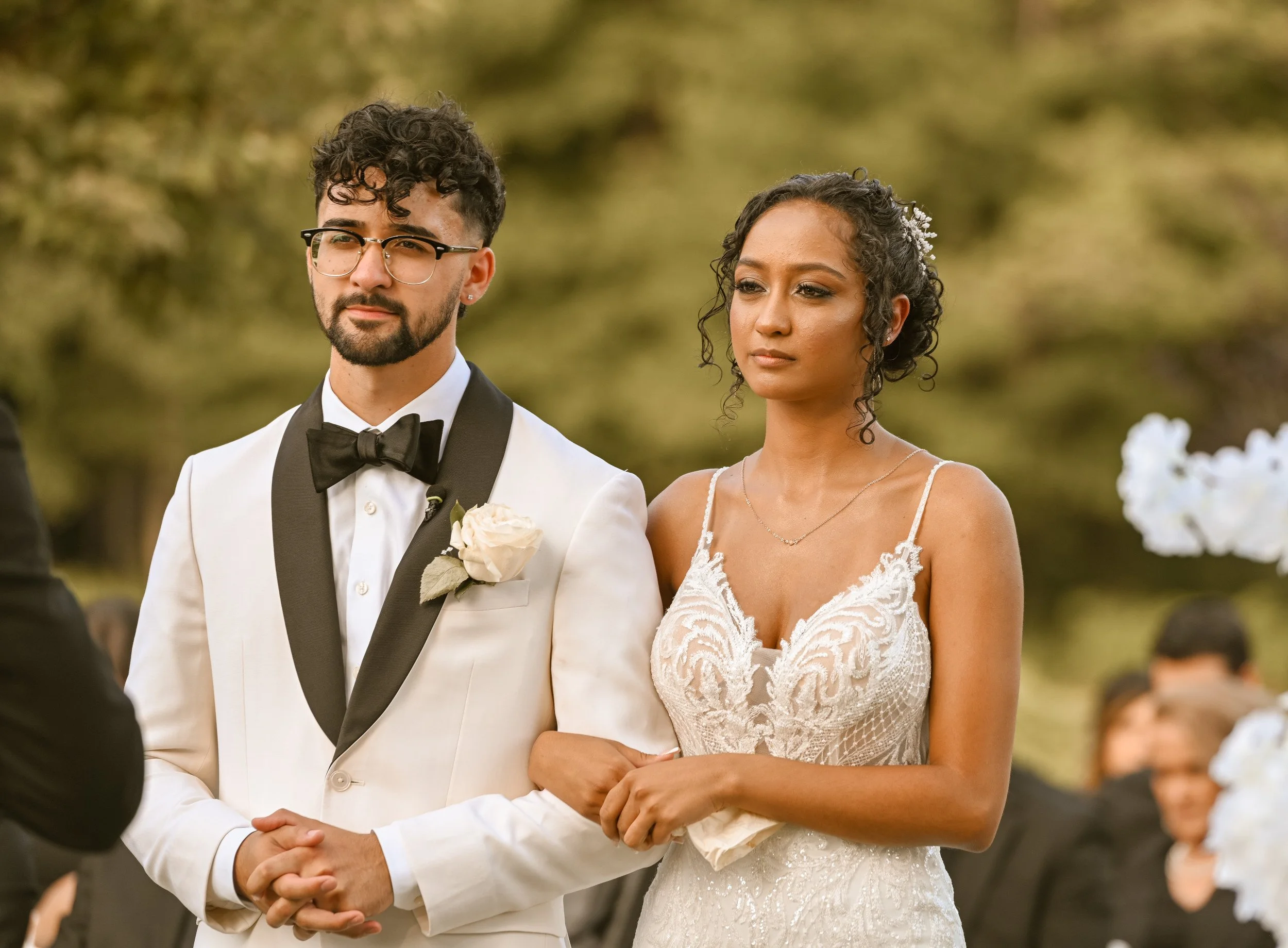 A couple getting married outdoors, with the groom in a white tuxedo with a black bowtie and glasses, and the bride in a lace white wedding dress, standing closely together during the ceremony.