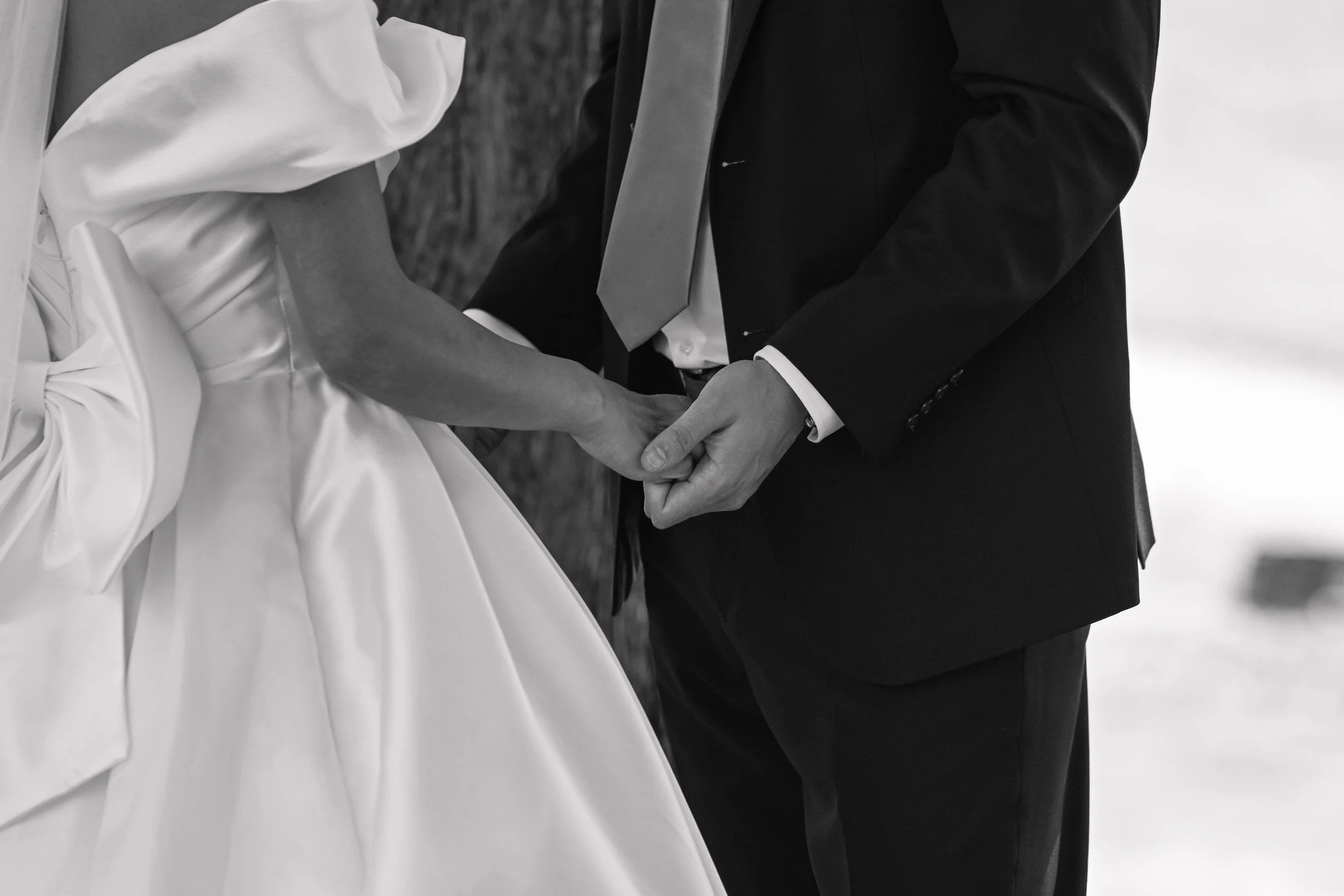 A couple holding hands during a wedding ceremony, with the bride in a satin gown and the groom in a dark suit, seen from the waist up.