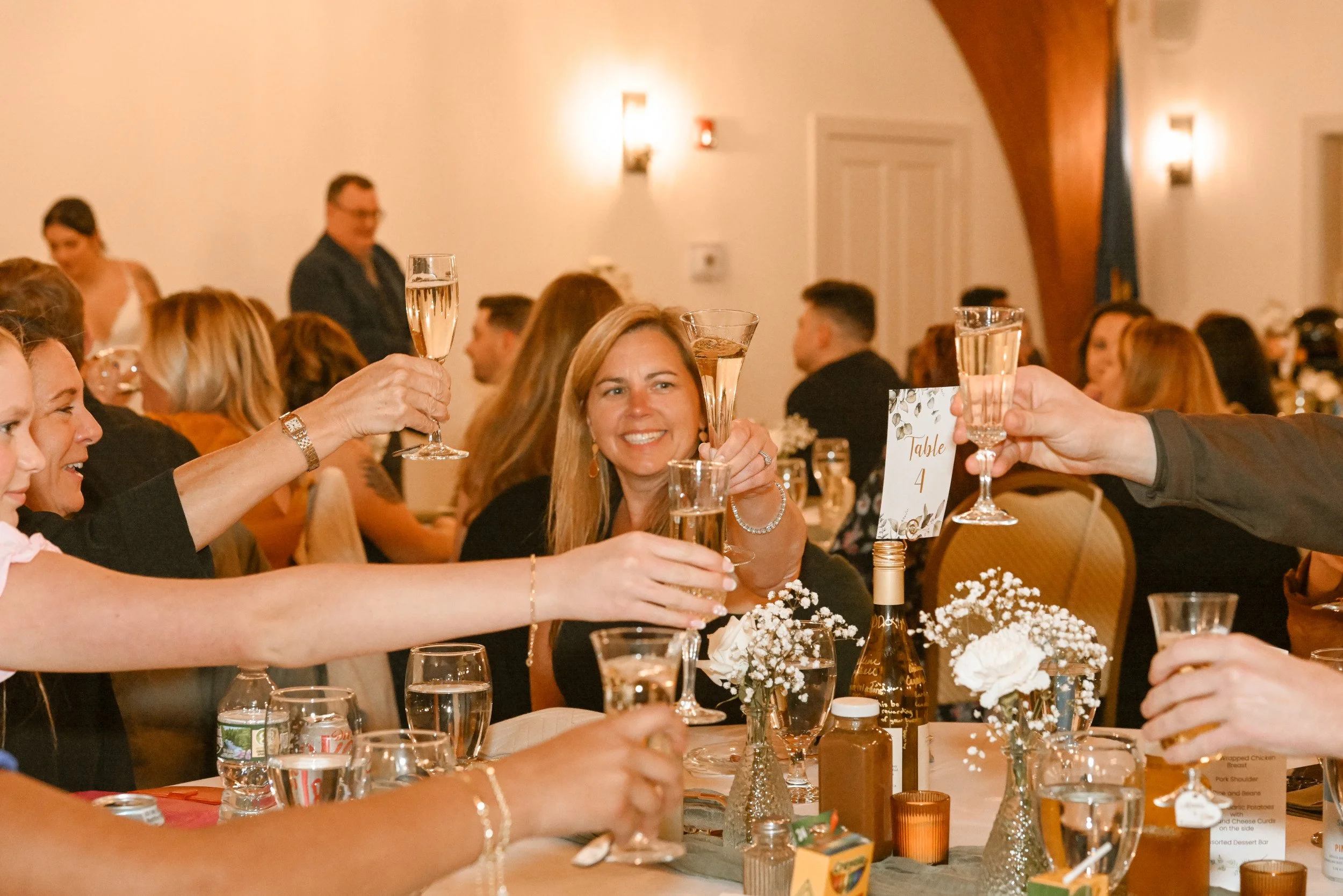 People at a celebratory gathering raising glasses for a toast, seated at a decorated table with flowers and drinks.