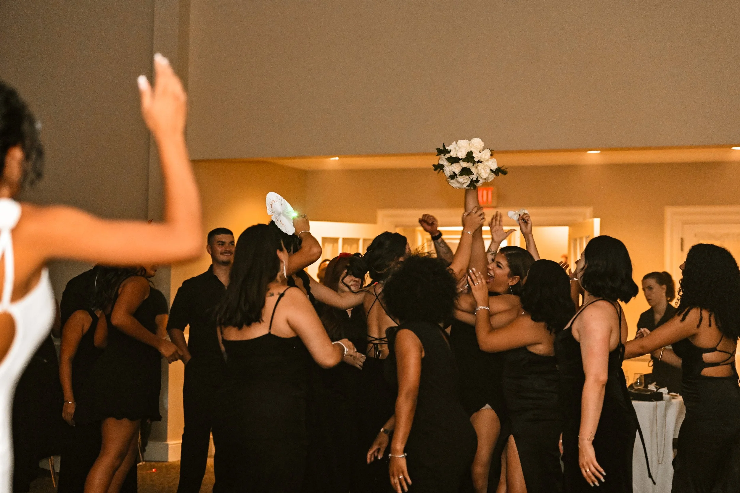 People dressed in black, dancing and celebrating at a wedding reception, with one person holding a bouquet of white flowers in the air.