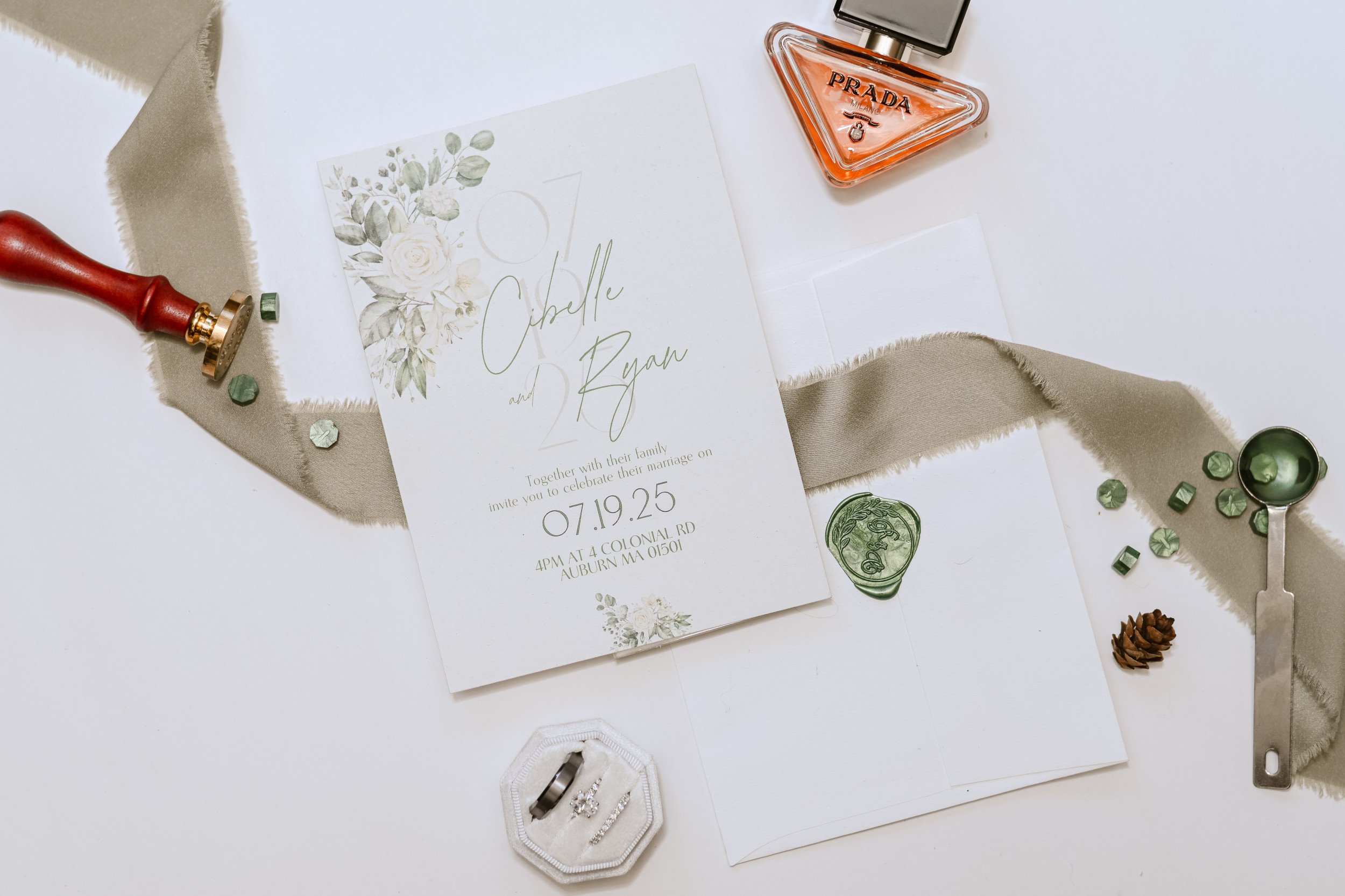 Wedding invitation card with floral design, perfume bottle, jewelry box with rings, decorative ribbon, pine cone, and scattered green sequins on a white surface.