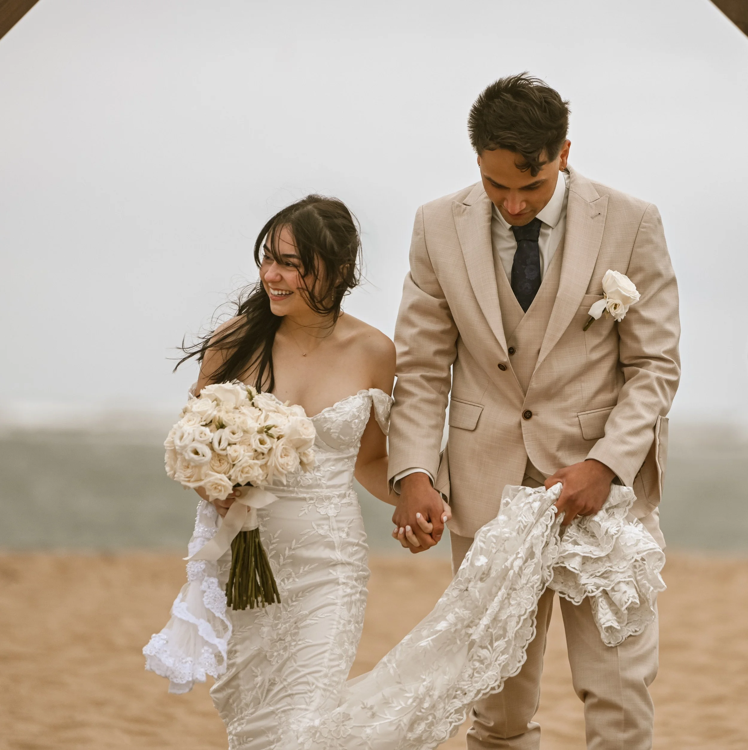 A bride and groom holding hands during their wedding ceremony. The bride is smiling and carrying a bouquet of white roses. The groom is dressed in a beige suit with a dark tie and a white rose boutonniere. They are standing in front of a plain white background.