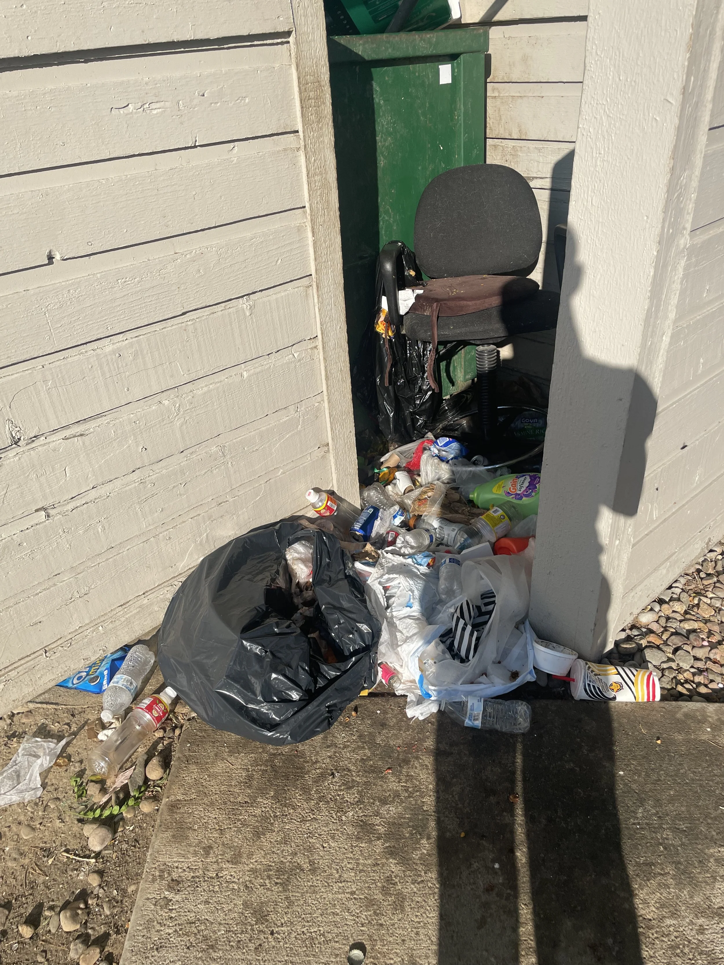 Overflowing trash spilling out of a black garbage bag onto a sidewalk next to a building wall, with discarded bottles and cups on the ground.