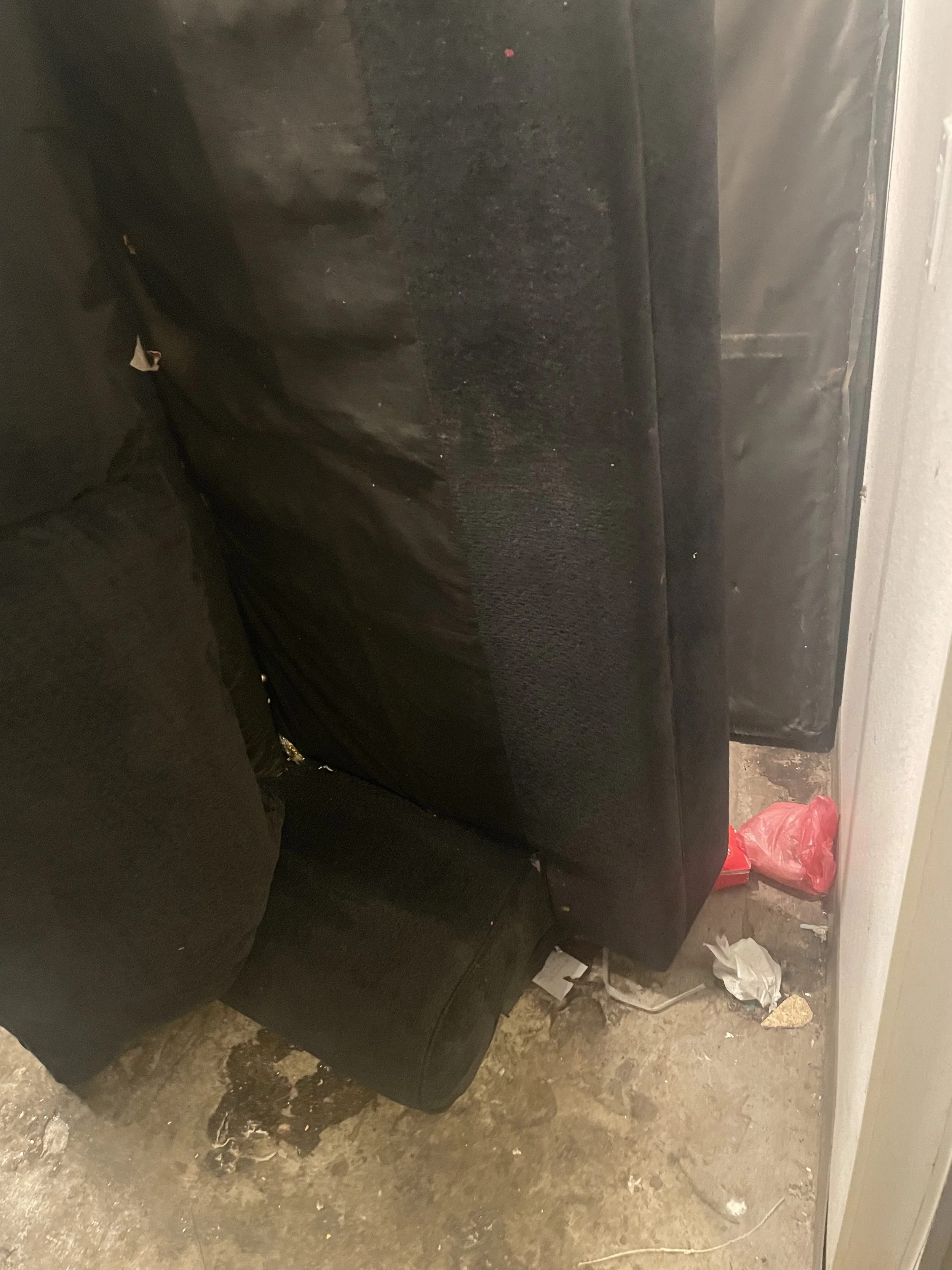 Cluttered space with a black couch, trash on the floor including a red plastic bag, crumpled paper, and a piece of cardboard, with a white wall and electrical outlet in the background.