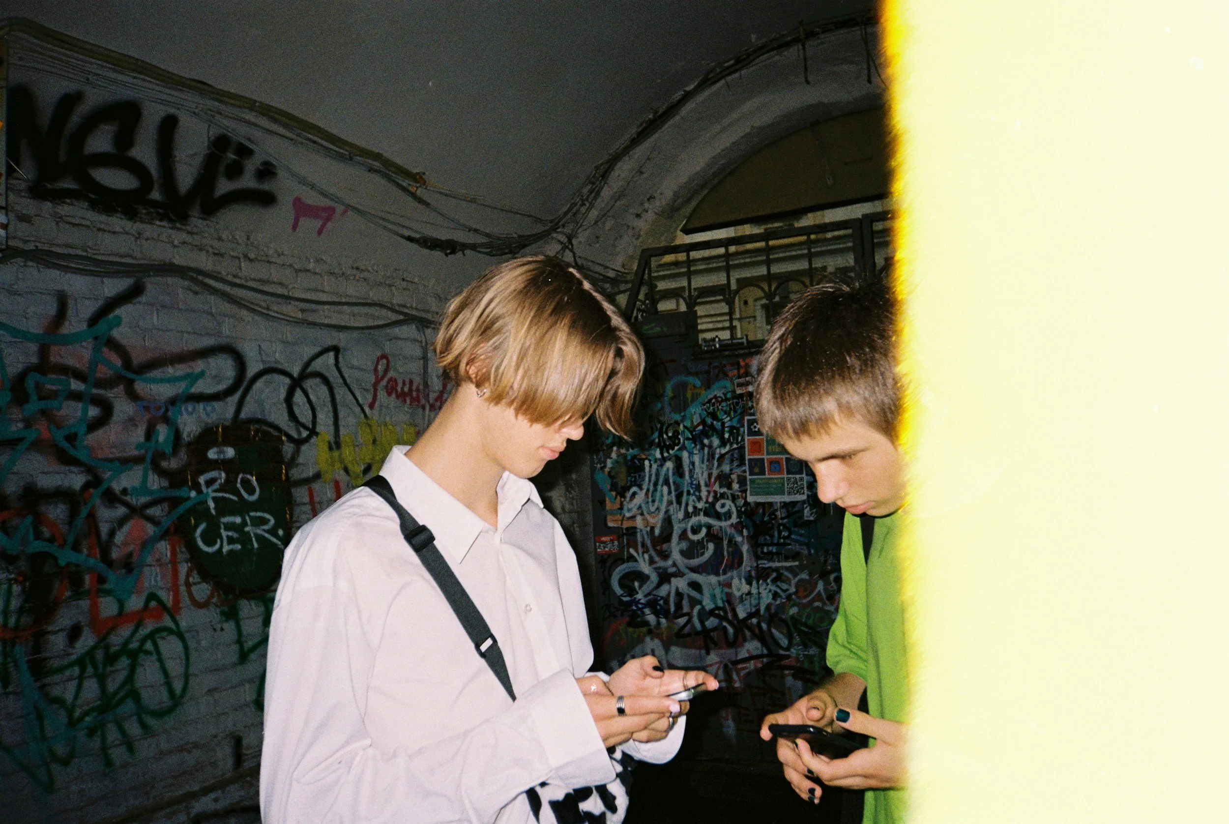 Two young people looking at their smartphones, standing in front of a graffiti-covered wall, with a yellow object partially blocking the right side of the image.