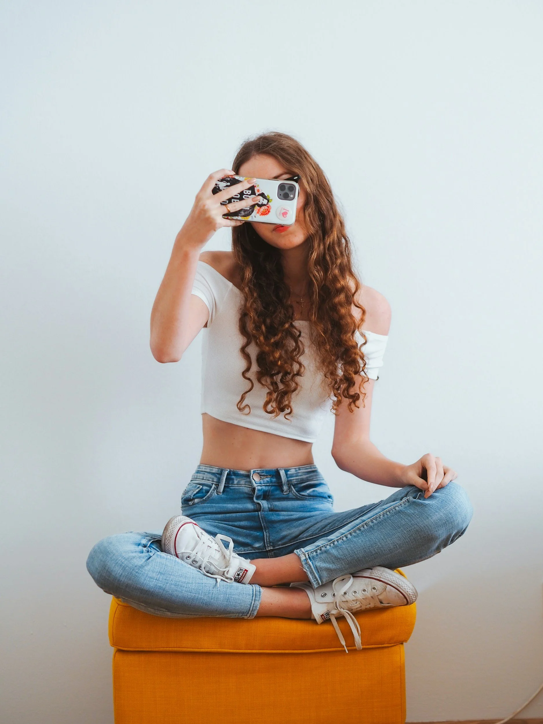 Young woman with long curly hair sitting cross-legged on an orange ottoman in front of a plain white wall. She is wearing a white off-the-shoulder crop top, blue jeans, and white sneakers. She is taking a selfie with her phone, covering part of her face.