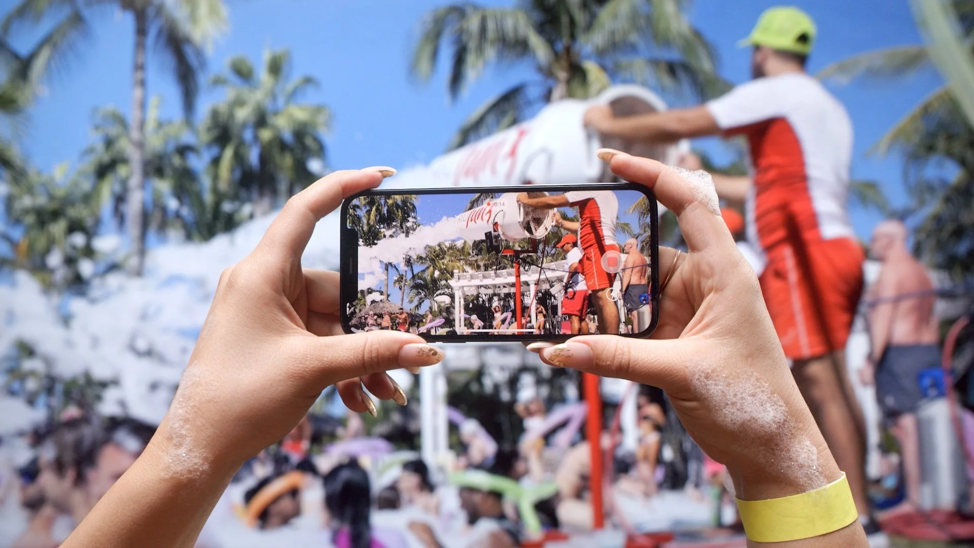 Person holding a smartphone taking a picture of a crowd participating in a water event outdoors, with palm trees and a clear blue sky in the background.
