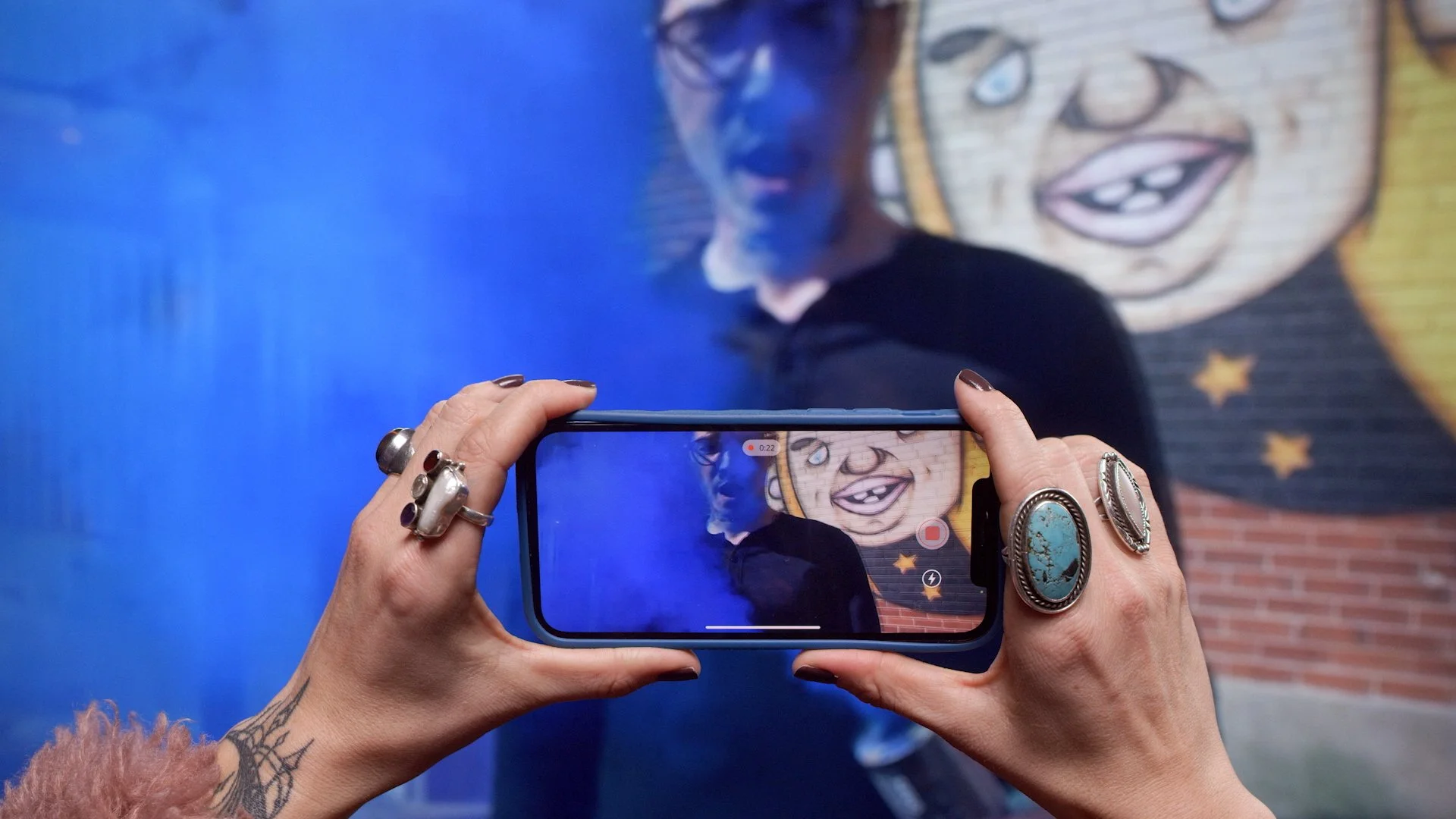 Person taking a photo of another person in front of a mural using a smartphone. The person in the background is smiling, and the mural features a cartoon face with a big smile, painted on a brick wall.