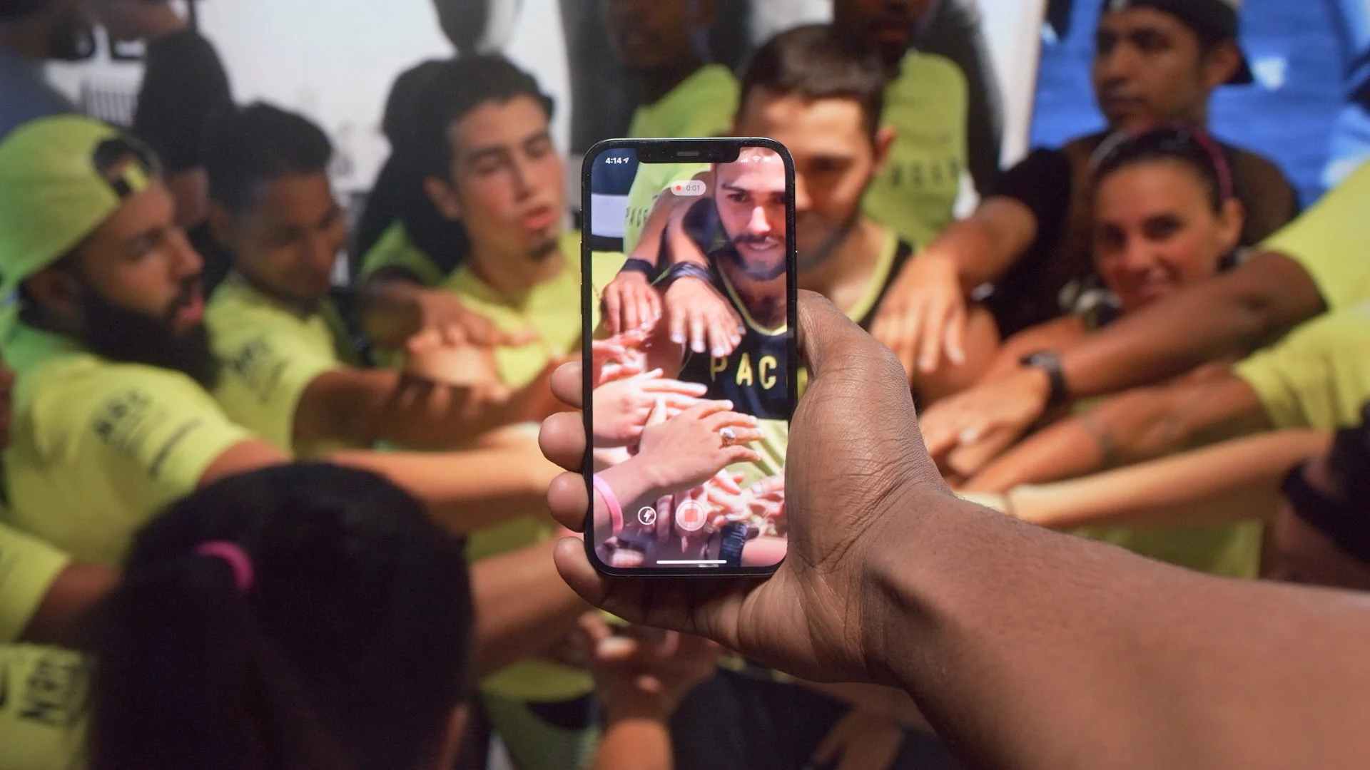 A person takes a photo of a group of people in a huddle, with all their hands stacked together in the center, using a smartphone.