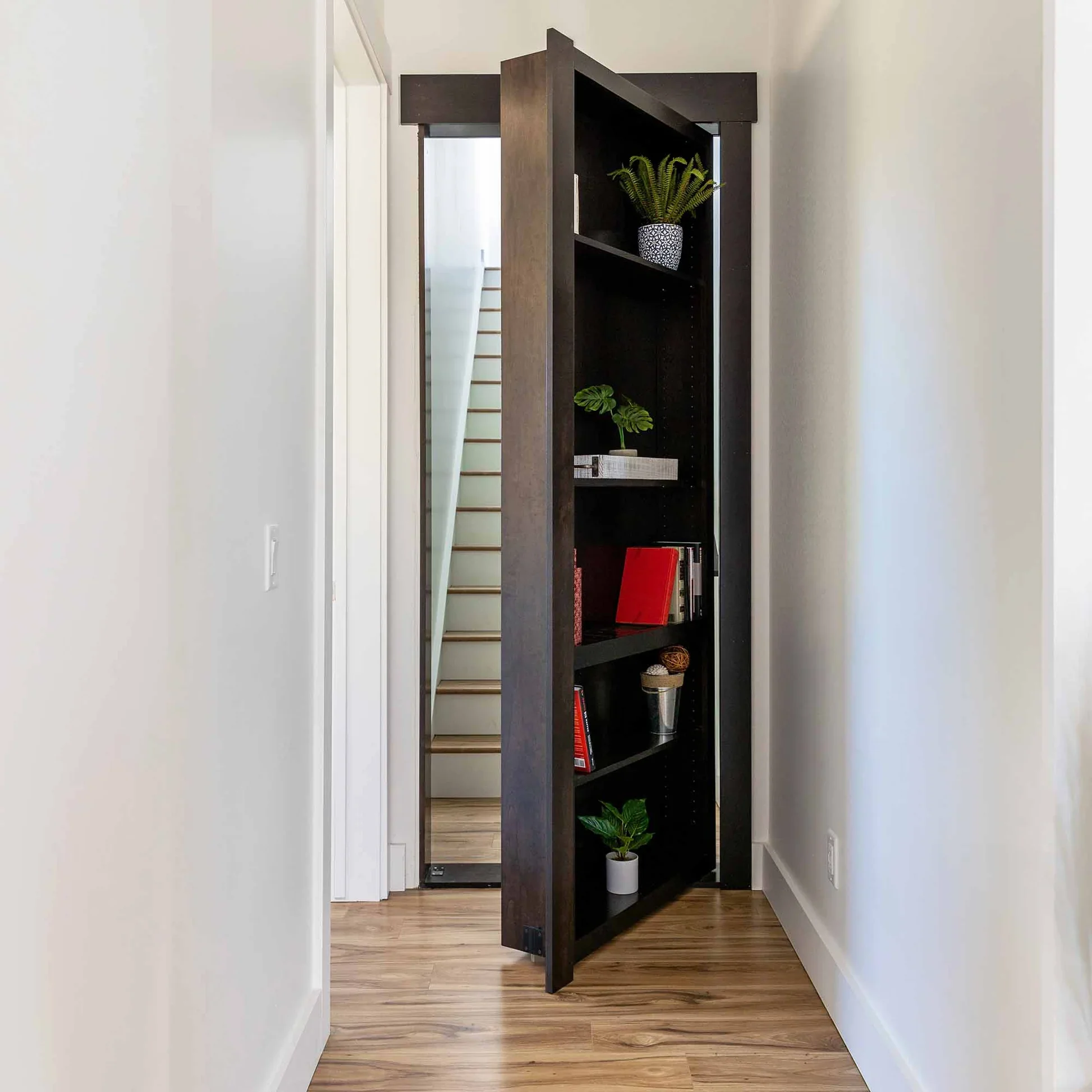 Black bookshelf with plants, books, and decorative items in a hallway with light-colored walls and wooden flooring.