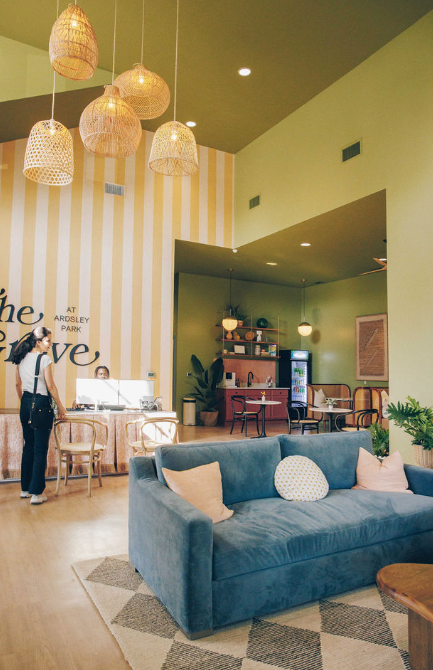Interior of a cozy, modern cafe with hanging wicker pendant lights, a blue sofa with pastel pillows, and a reception desk. A woman checks in at the desk, and the decor features green and striped walls with plants and wooden furniture.