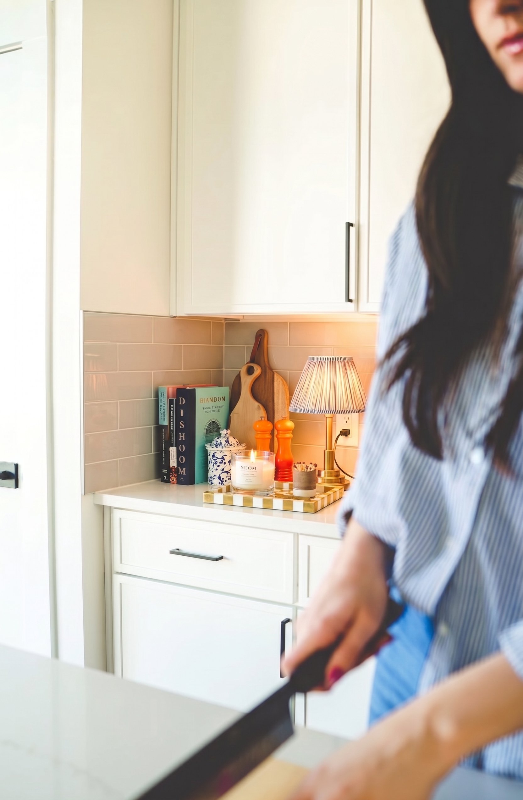 Close-up of a person in a striped shirt with long dark hair, preparing food in a modern kitchen with beige cabinets, a white countertop, decorative items, and a lit table lamp.