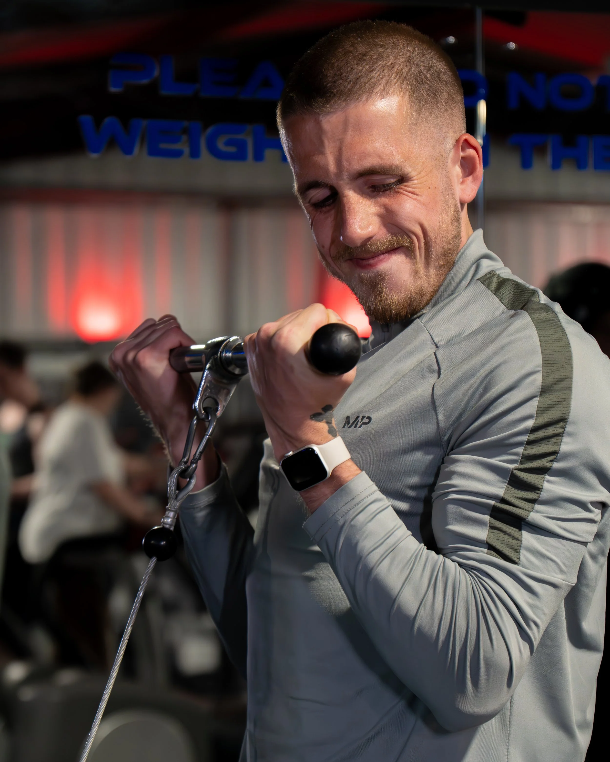 Smiling man with short hair and beard holding bike handlebar, wearing a gray sports shirt and smartwatch, in a gym or fitness center.