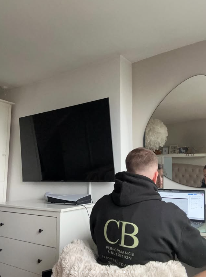 A person working on a laptop in a room with a large wall-mounted TV, a round mirror, and a white dresser. The person is wearing a black hoodie with green lettering "CB" and some text underneath.
