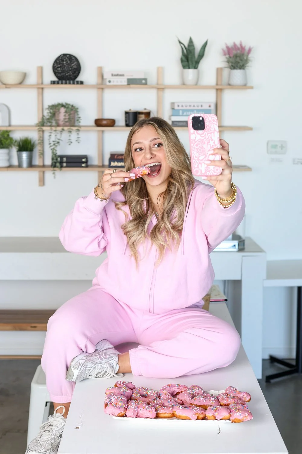 woman in pink holding a donut about to eat it and a plate in front of her, taking a selfie with donut in hand, sitting on a counter infront of a bookshelp