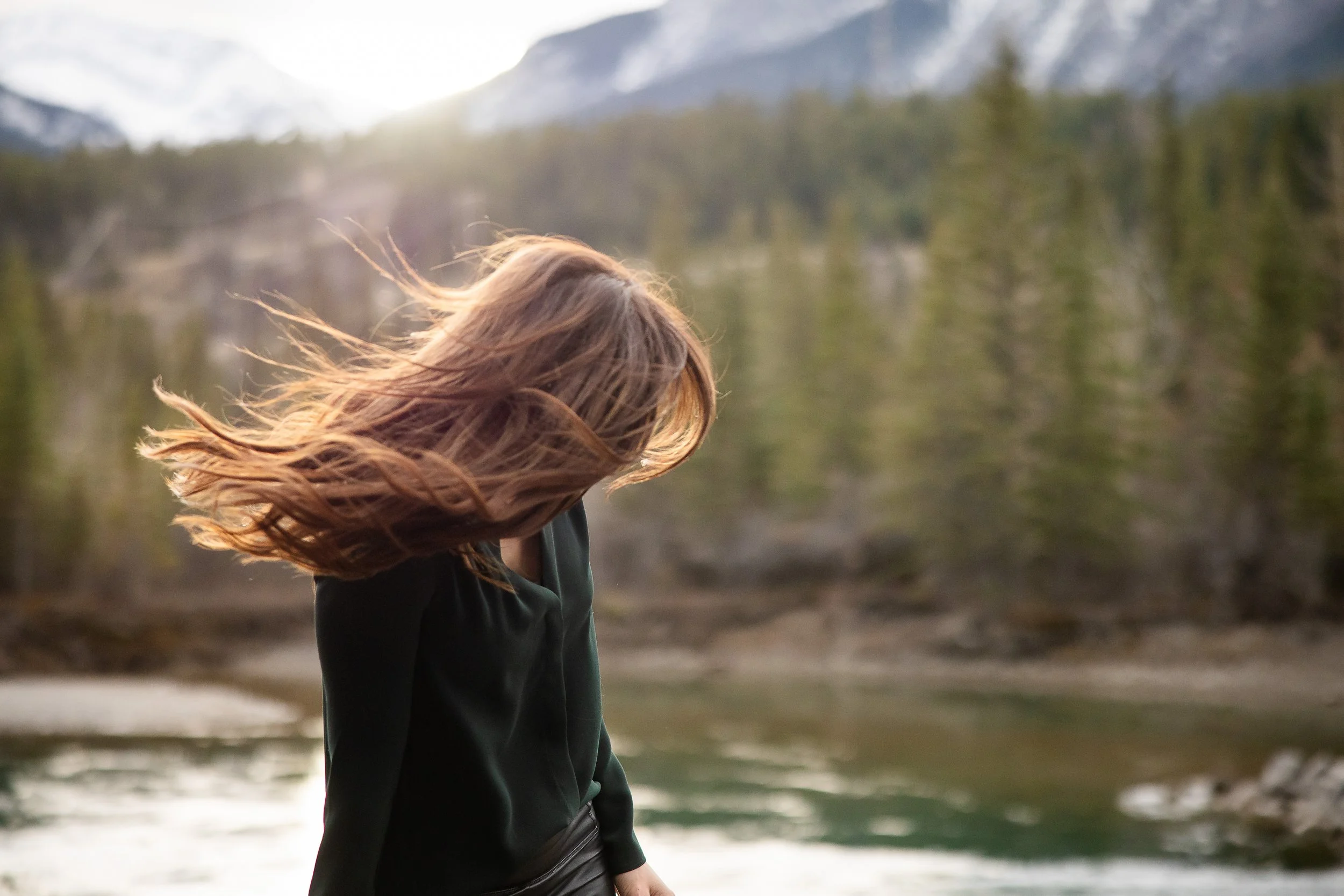 A woman with long, wavy hair standing outdoors near a river, with a forest and mountains in the background, during sunset or sunrise.