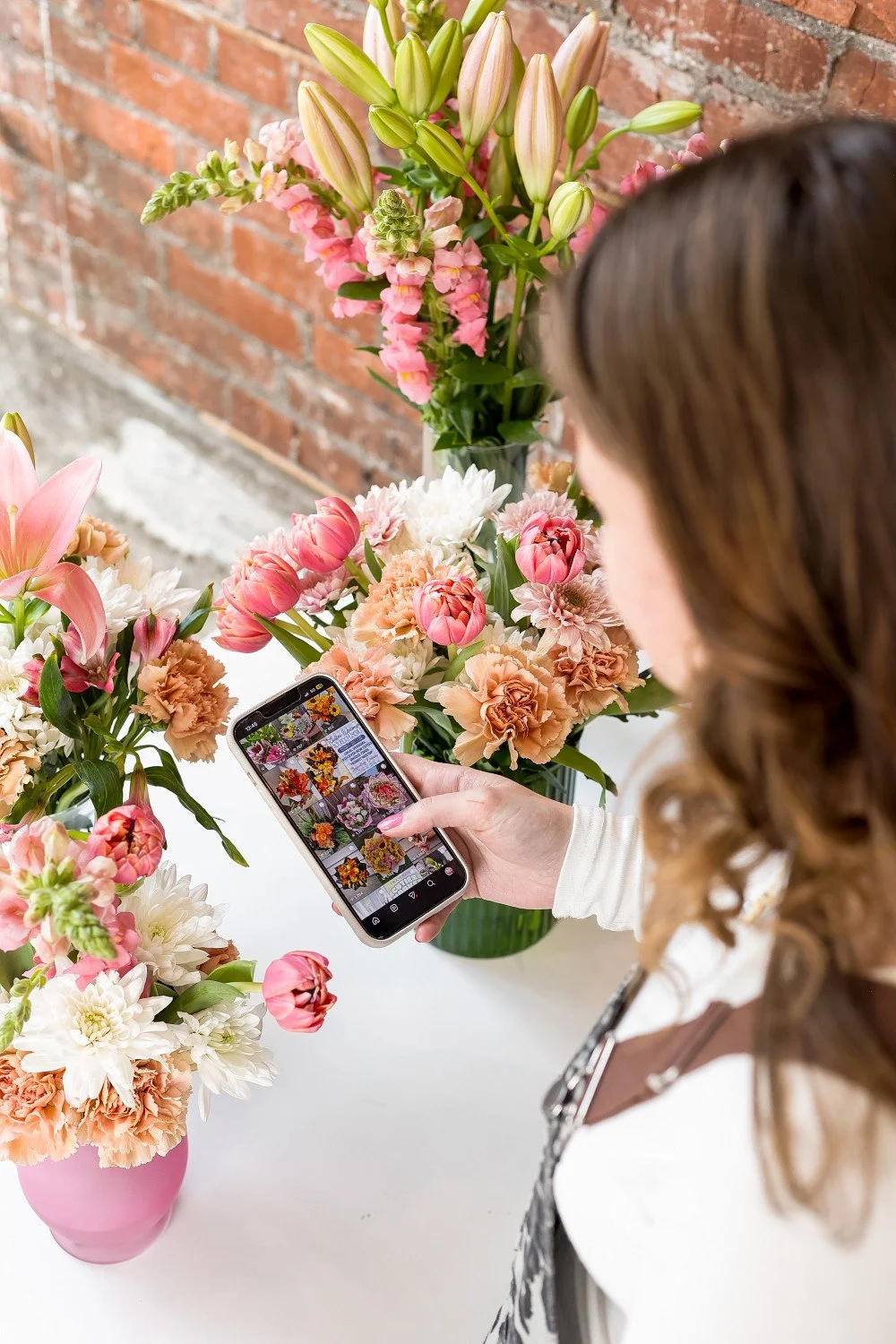 image of woman holding her phone with instagram profiles pulled up and a lot of florals in the fram, brick wall can be seen on the left side, woman is wearing overalls and has pink finger nails