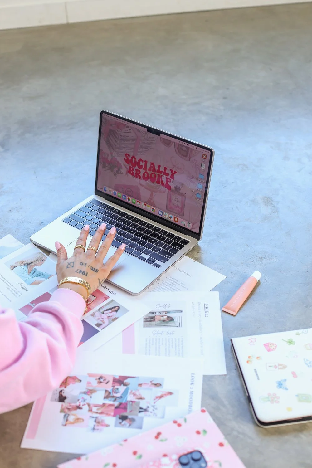 woman with hand on laptop showing her logo, papers on front and lipgoss in sight, laying on floor with tatoos on her hand