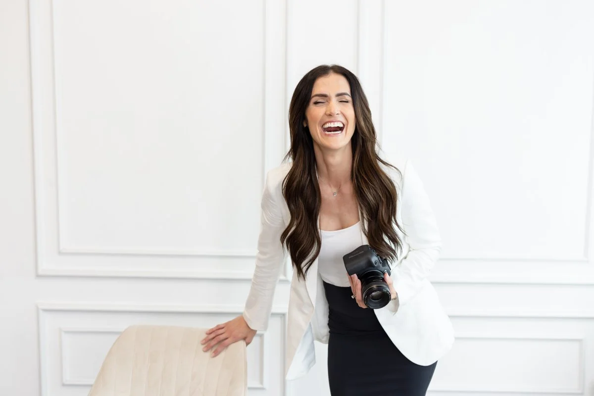 A woman in a white blazer and black skirt smiling and holding a camera, standing in a bright room with white walls.