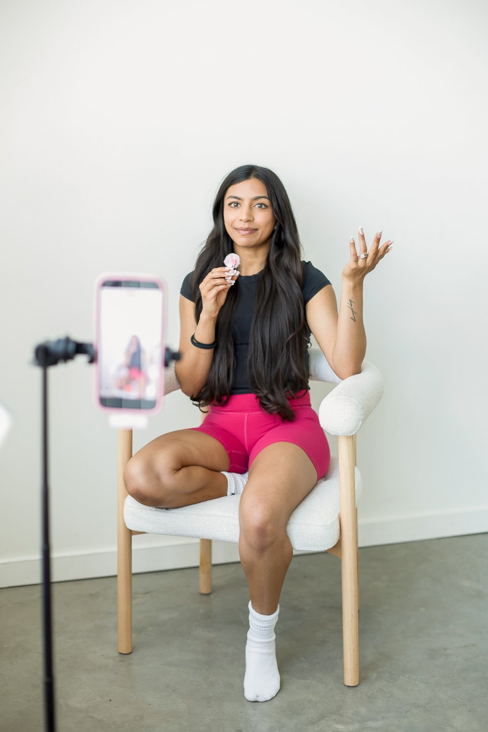 woman in pink shorts holding a pink microphone with a phone camera in front taking a video on a white chair
