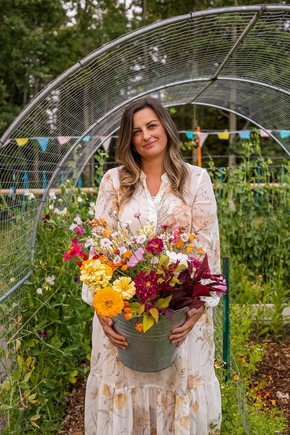 woman holding a bucket full of flowers wearing a dress with smile on her face in front of her garden