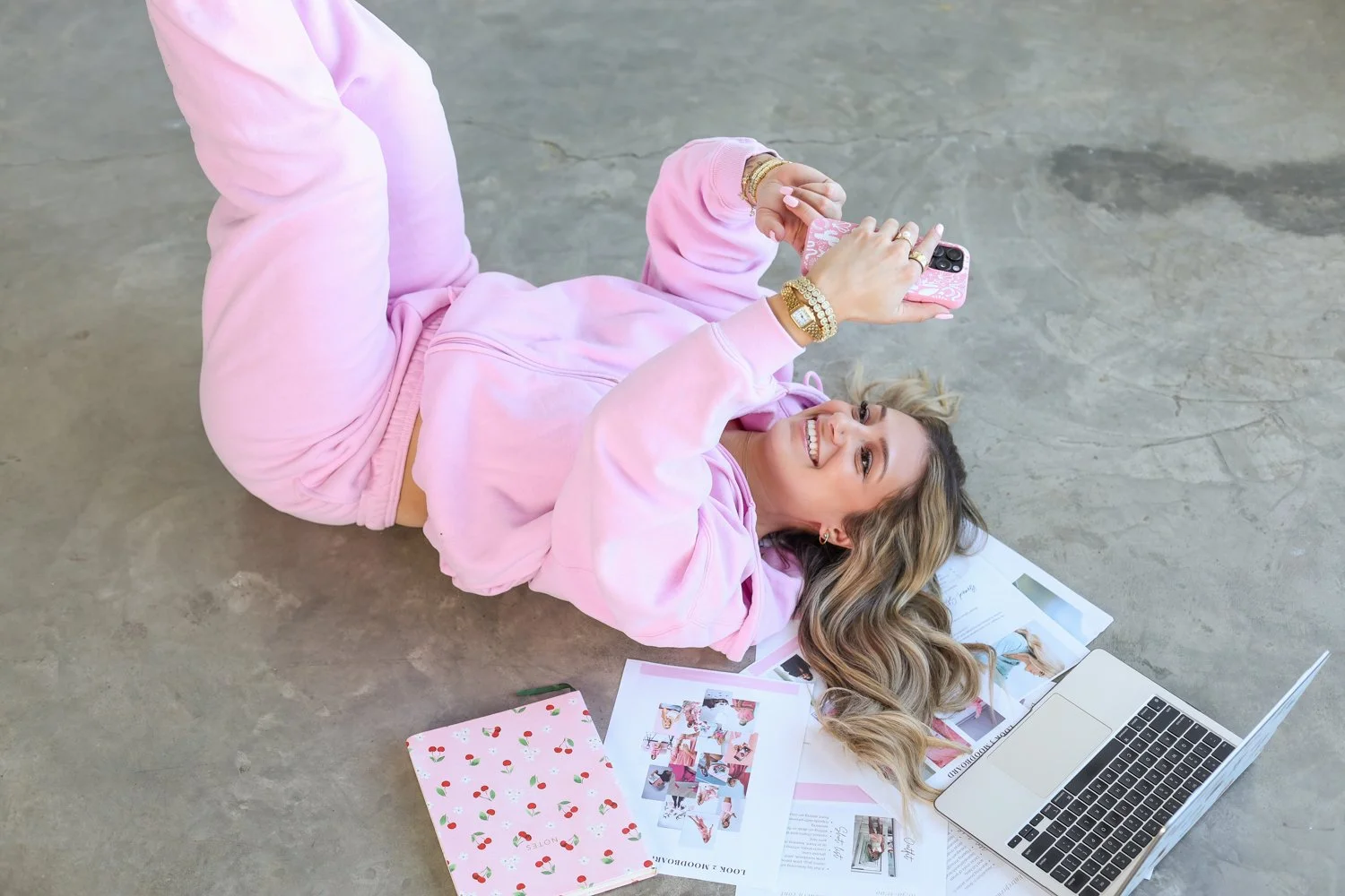 woman laying on back holding phone infront and smiling with notebook and papers and laptop ont he floor, blonde hair and jewellery