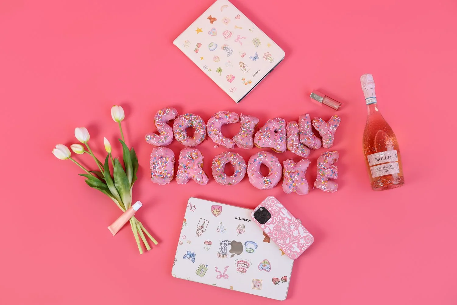 donuts spelling socially brooke with tulips and prosecco and laptop with phone and lip gloss and a notebook