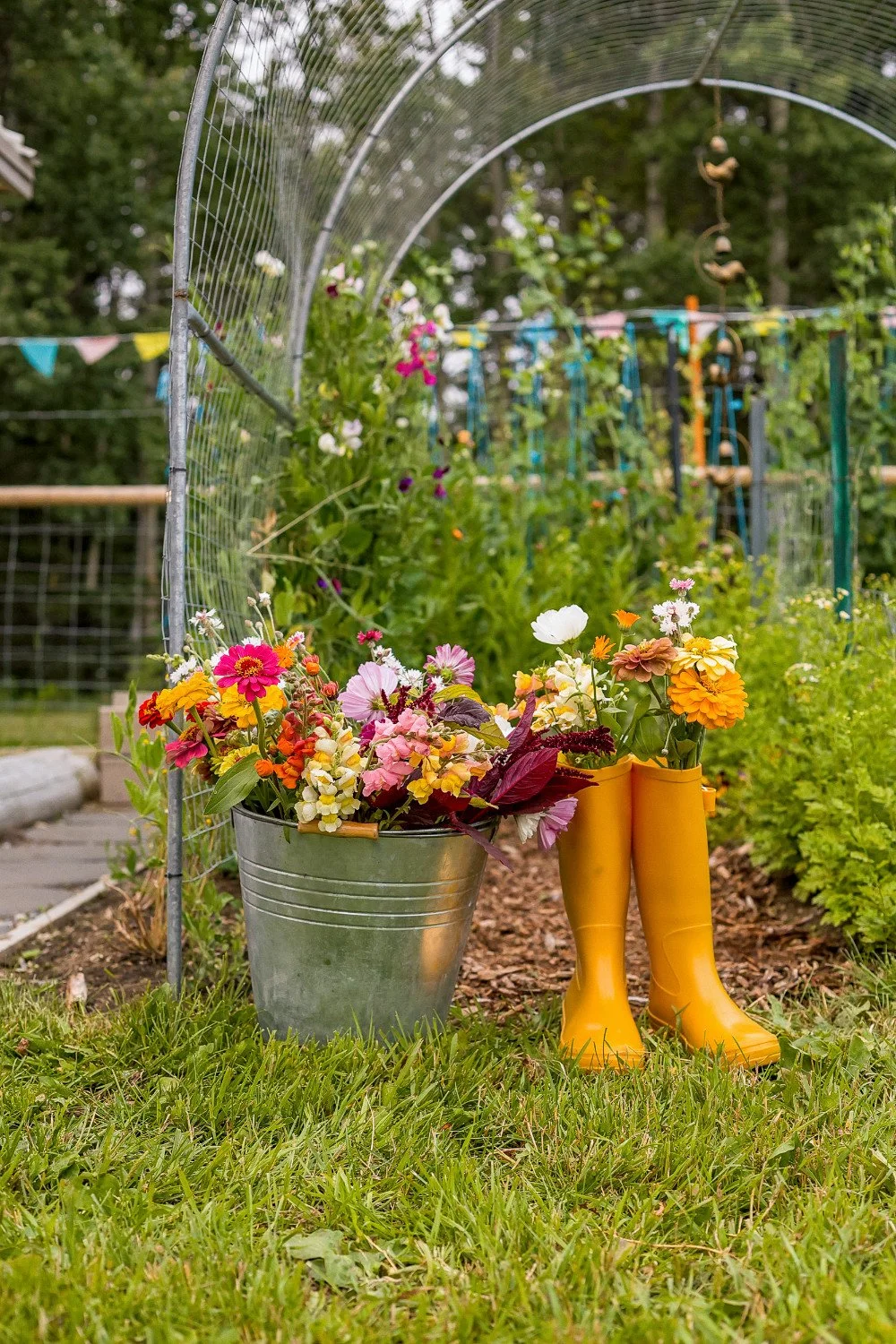 phtoo of flowers in yellow rubber boots and a bucket full of more flowers in the middle of a garden field