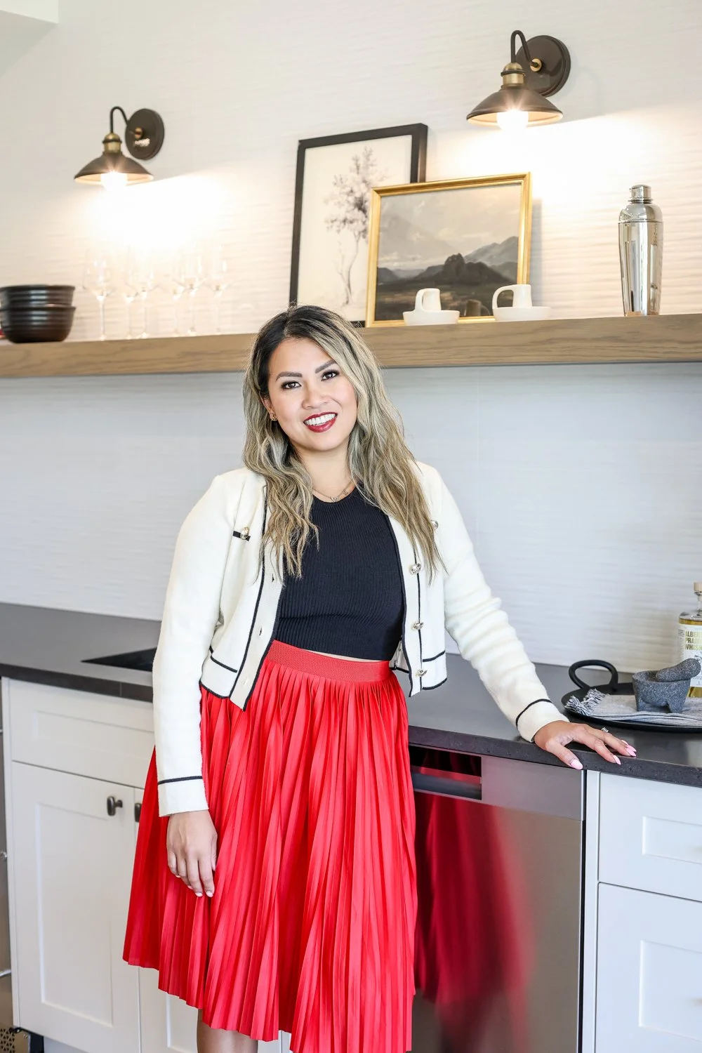 woman standing in kitchen looking at camera with smile on her face, red skirt and hand on counter