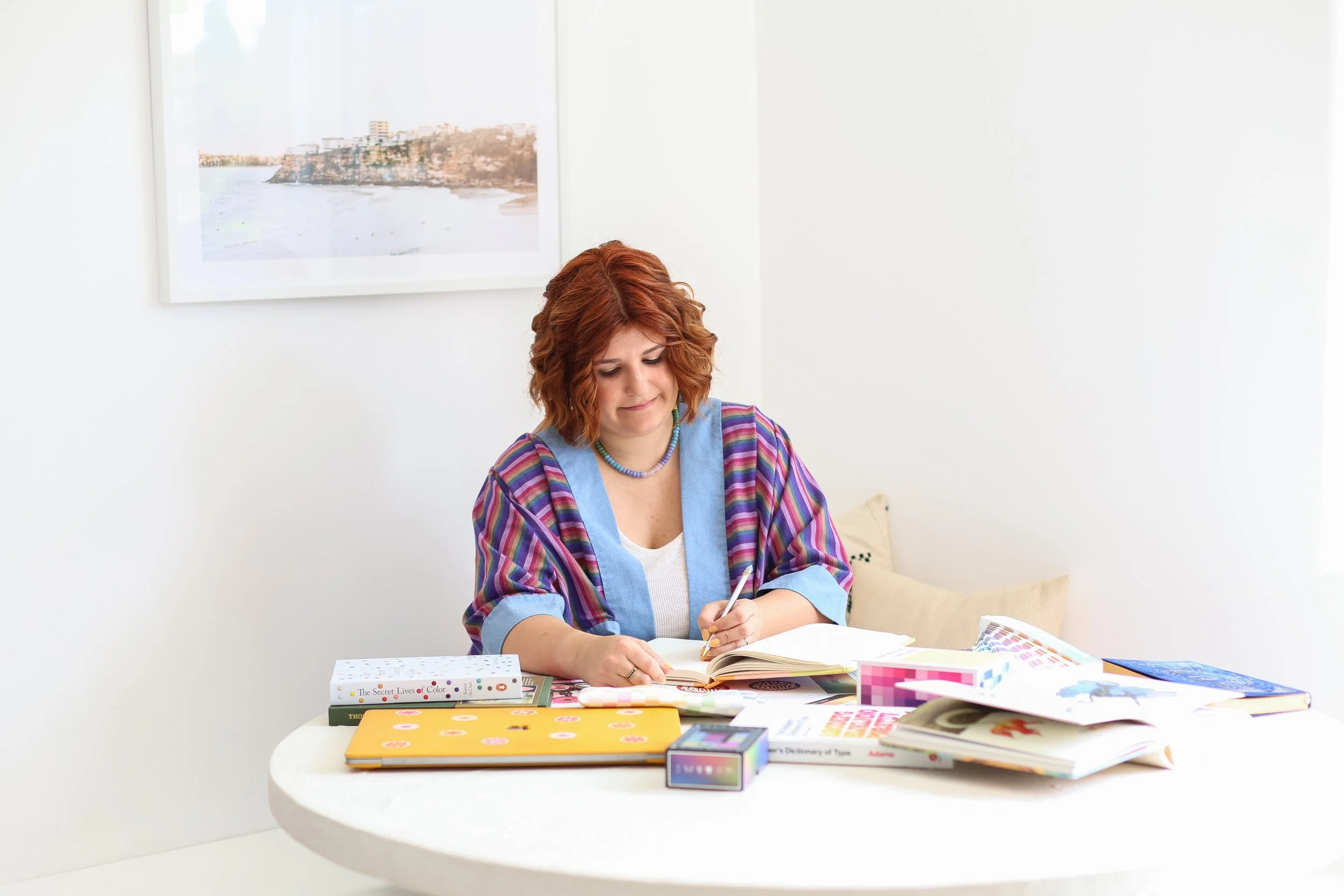 woman in colourful top with books layed out on table, looking down with pen in hand writing