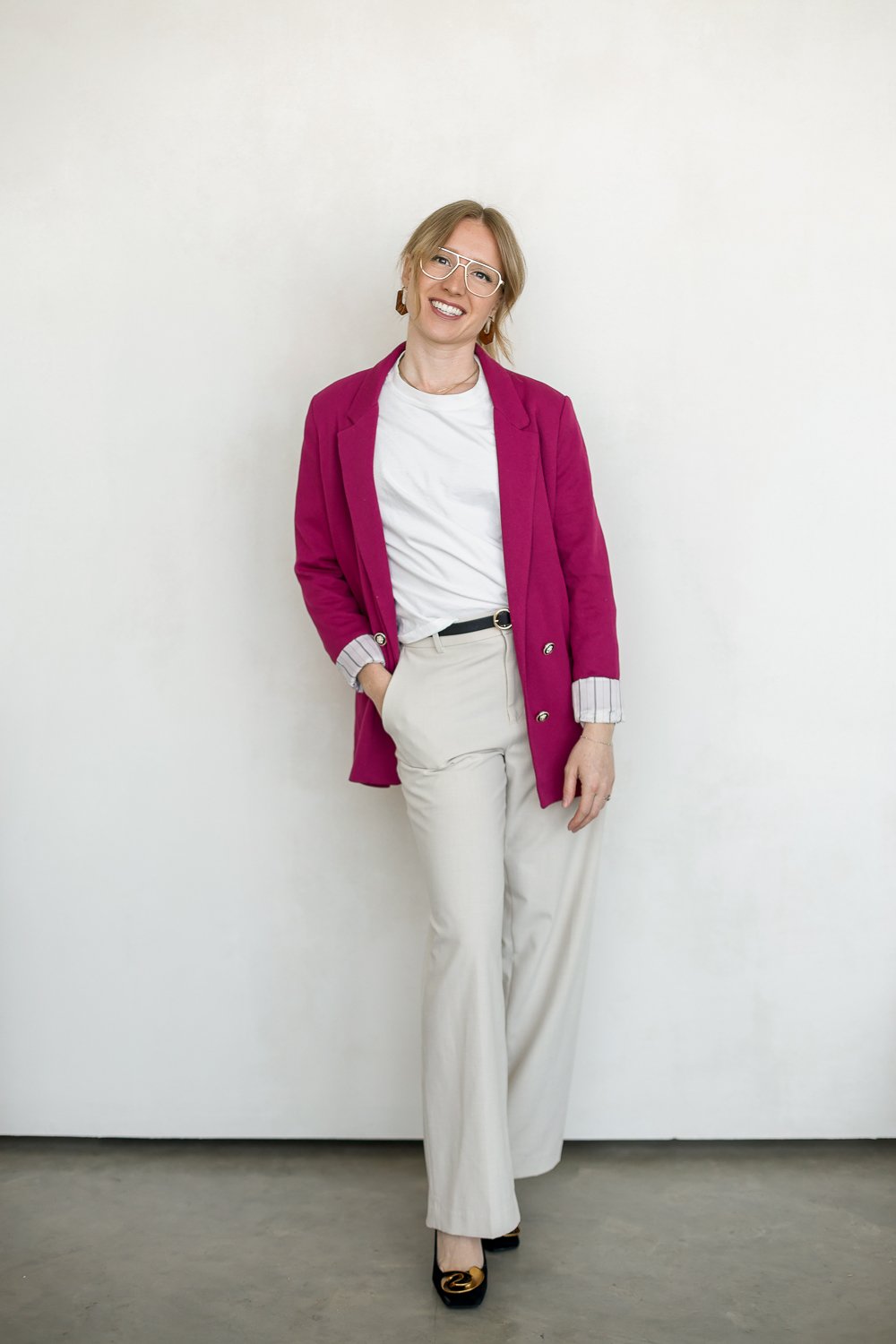 woman with glasses and pink blazer standing smiling at camera one hand in pants pocket