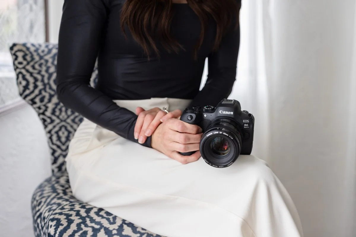 A woman with long dark hair sitting on a patterned chair, holding a Canon EOS R6 camera with a lens, in a well-lit room.