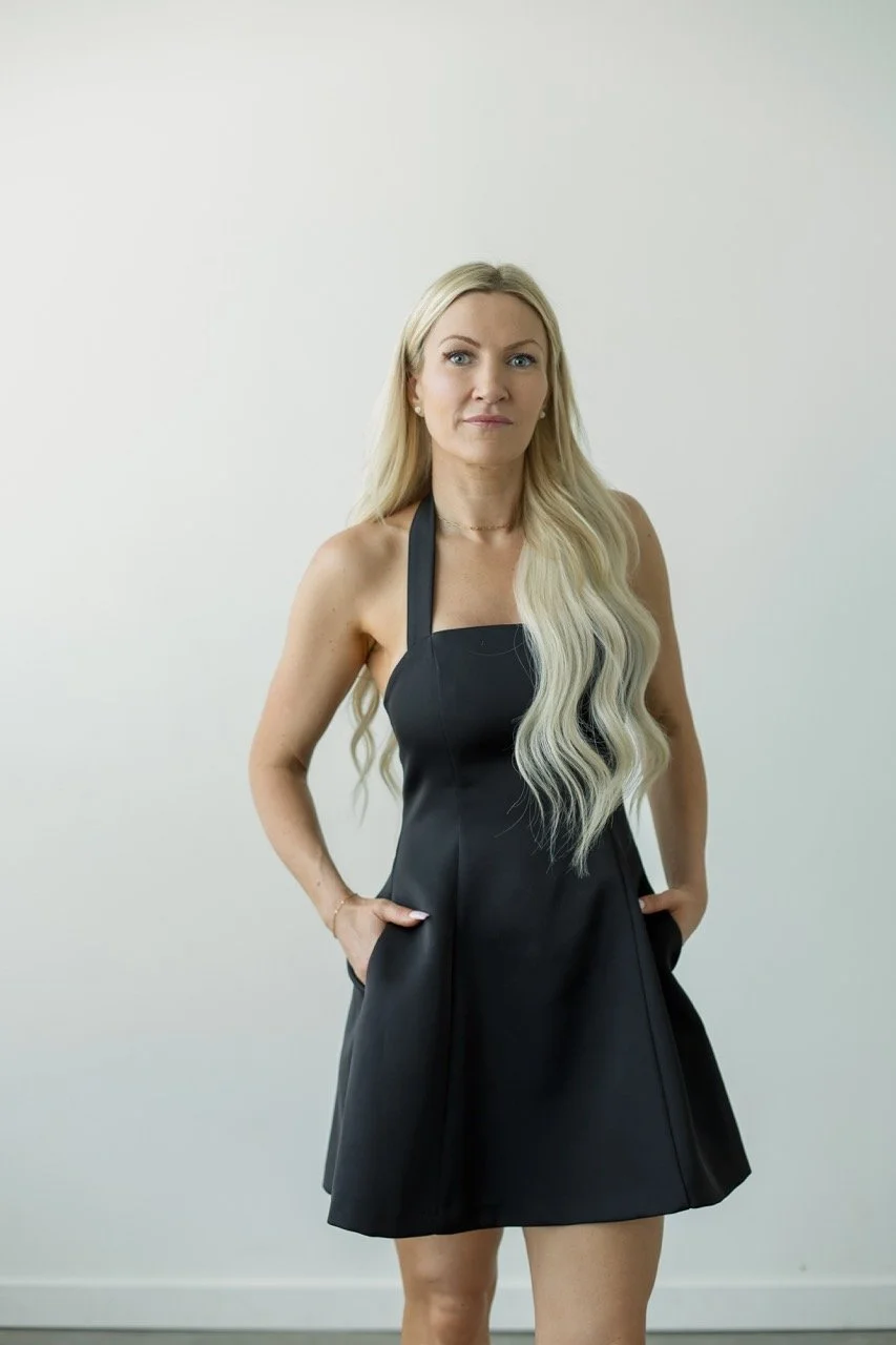 woman with long blone hair and black dress with hands in pockets looking at camera
