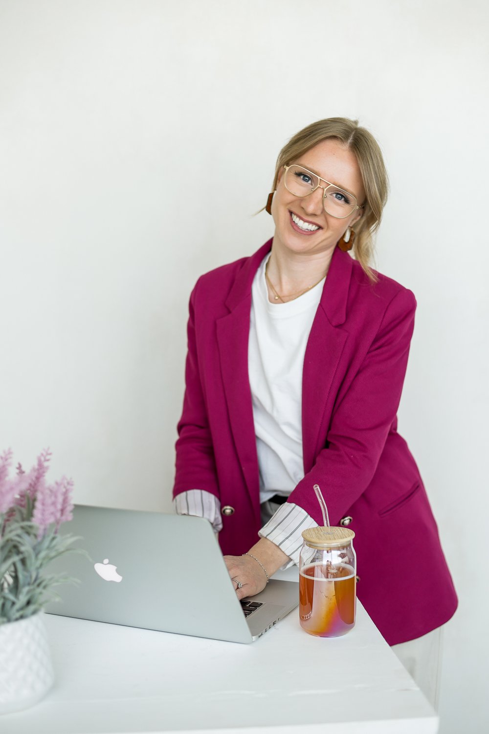 woman smiling at camera with pink blazer and glassed, compuer in front and a glass and flowers in the scene