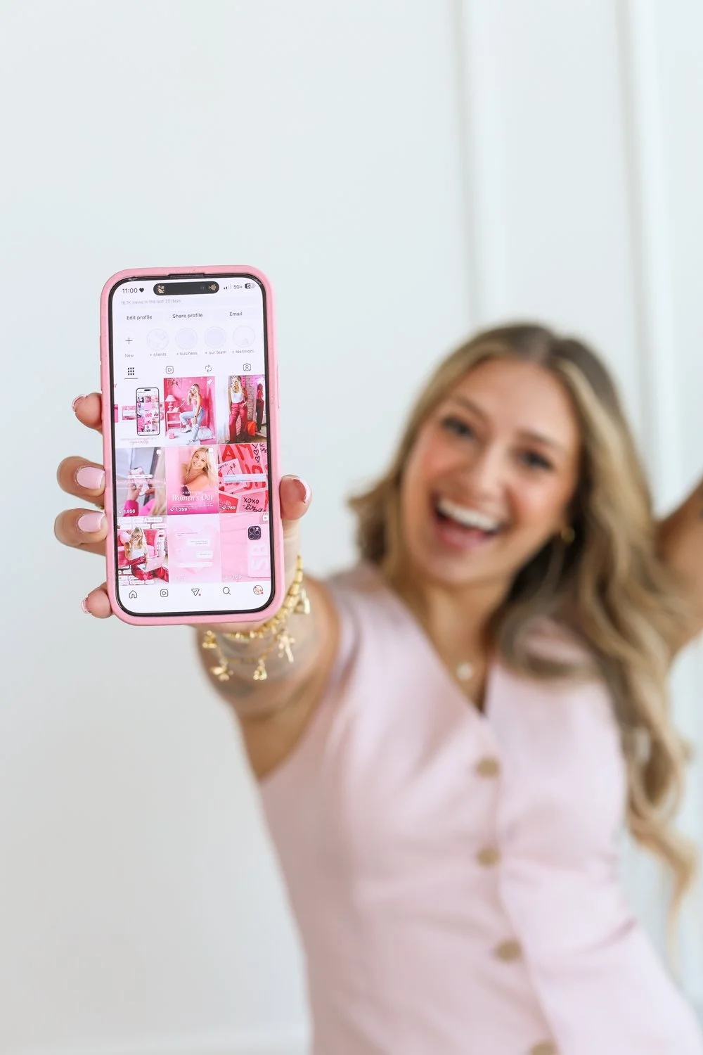 woman holding phone out in front showing her instragram feed, smile on her face wearing pink shirt and lots of jewellery showing