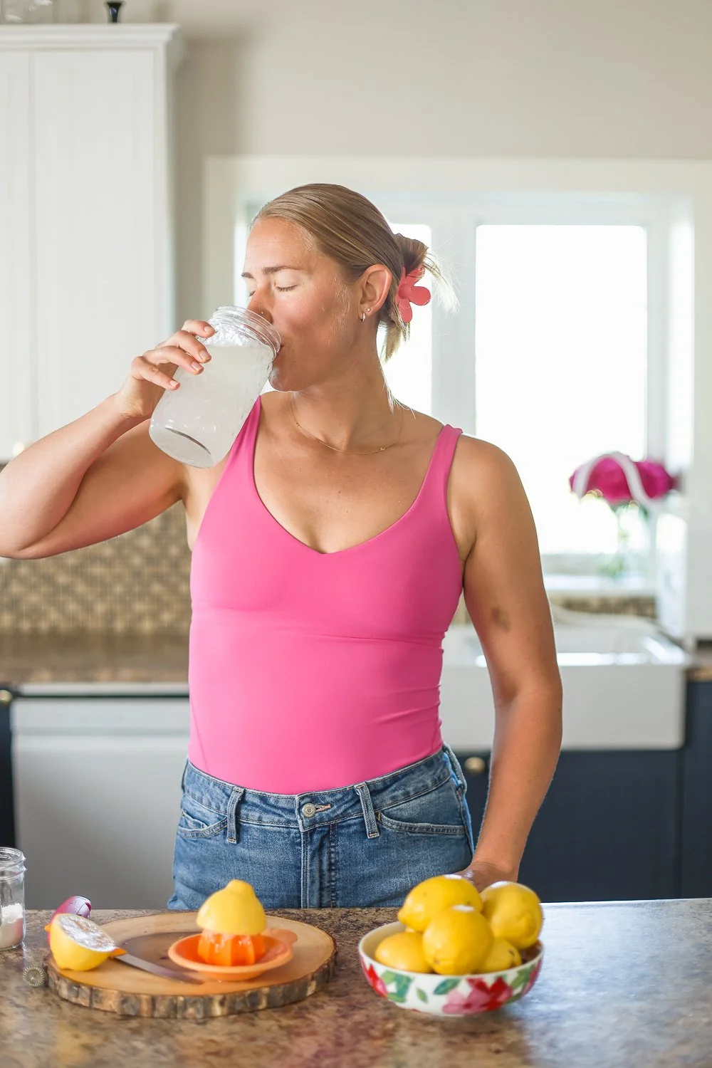 woman drinking a glass of water in a pink shirt at her counter with lemons on the counter and her eyes are closed.  Pink flowers in the background in front of the window