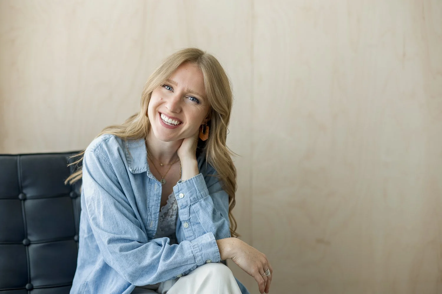 woman with hand behind her neck, sitting on black chair leaning forward in denim collared top and white pants