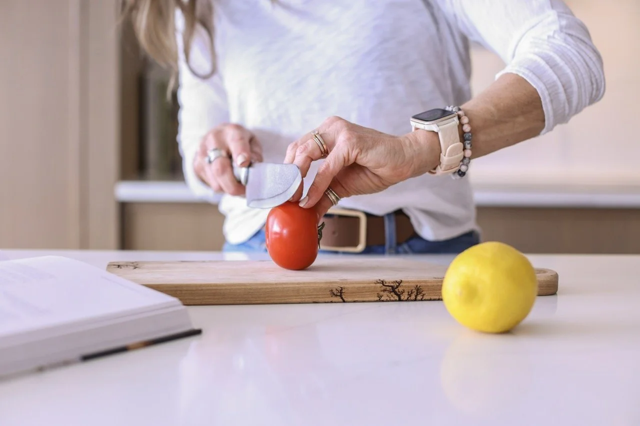 woman in white shrt cuting a tomato on a wooden cutting board with lemon and cookbook in the forground