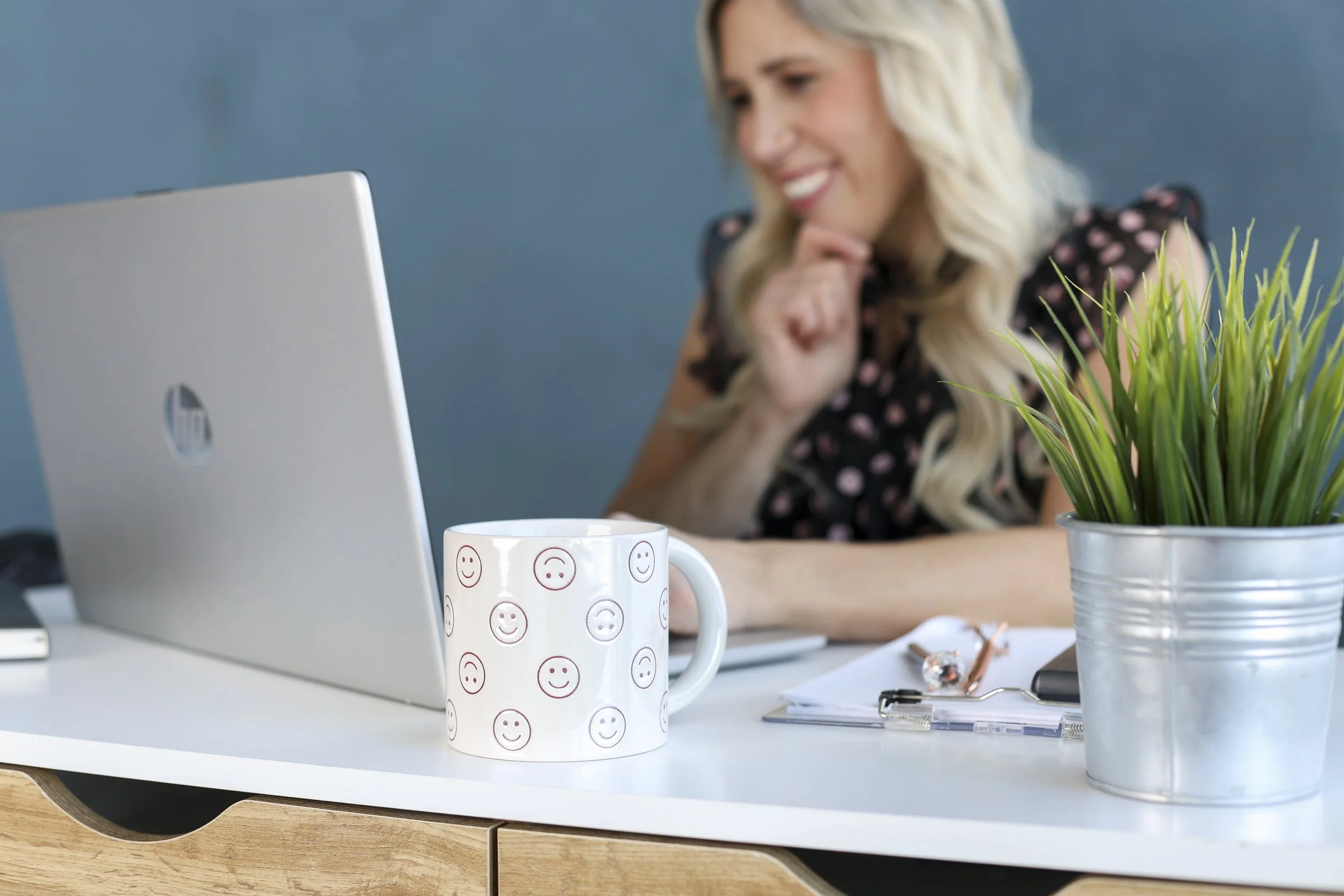 woman looking at computer ith mug in forground and a clipboard with pen and green plant in front with blue wall behind