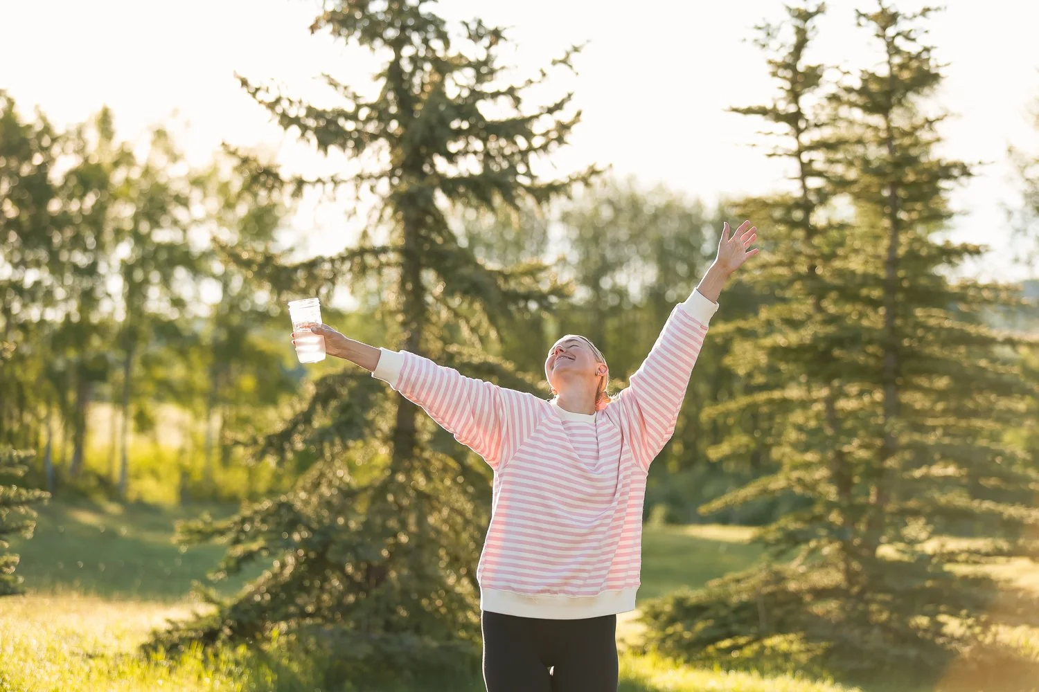 woman holding a glass of water with arms raised and a smile on her face in front of a row of trees