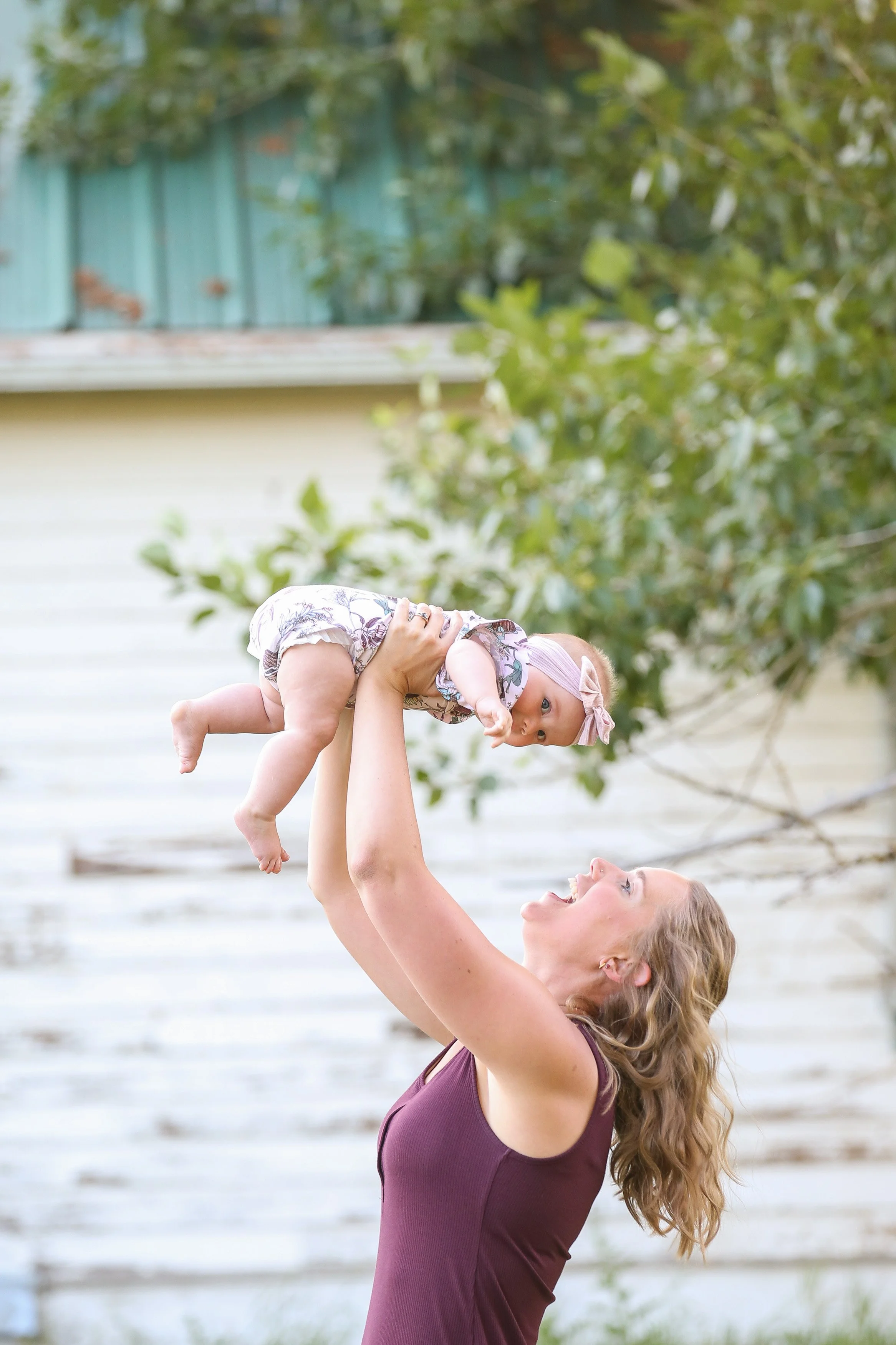 A woman with wavy blonde hair lifting a baby girl into the air outdoors. The baby is wearing a light pink headband and a floral outfit, and the woman is smiling while looking up at the baby. There are trees and a house in the background.