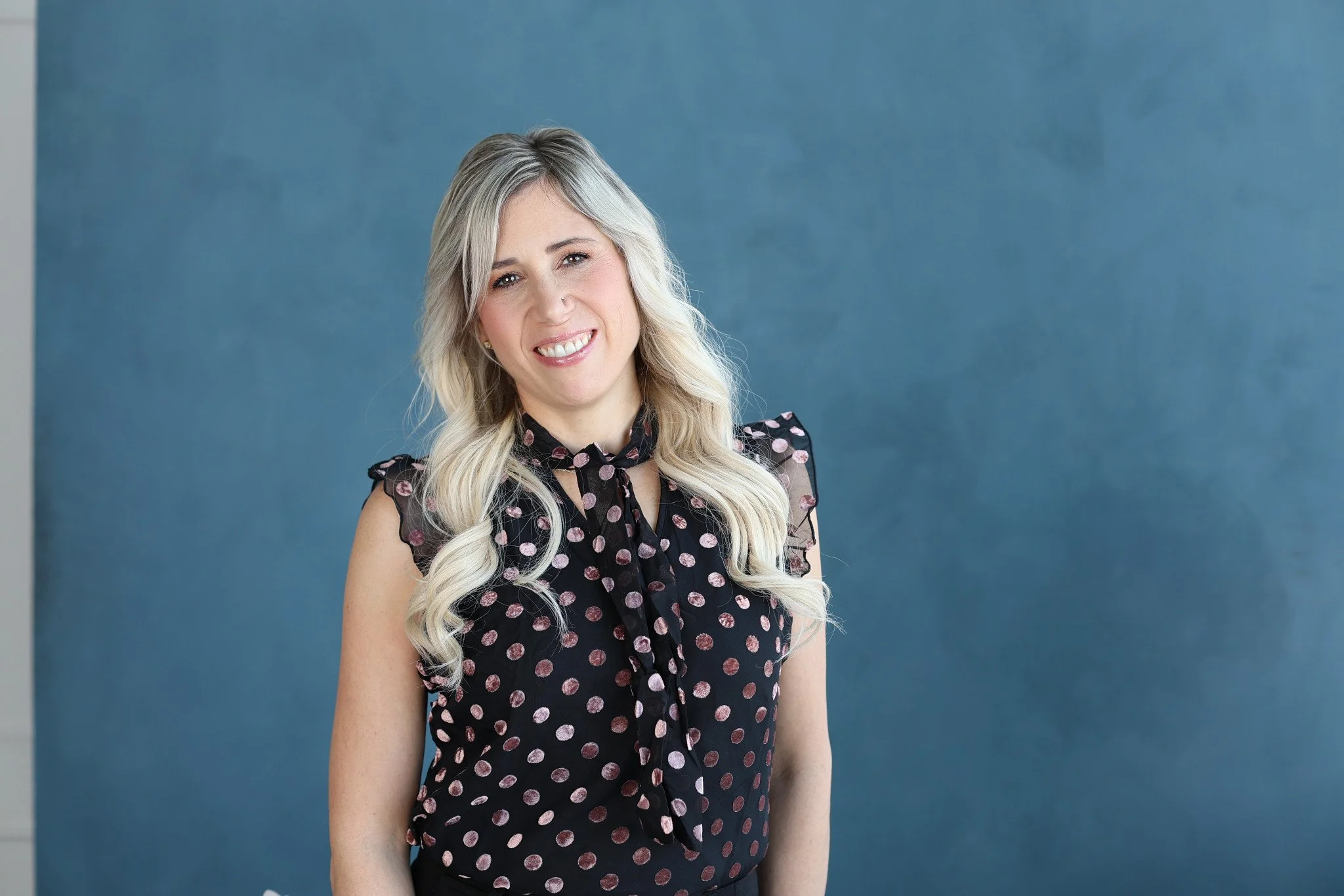 Smiling woman with blonde hair in loose waves, wearing a black blouse with pink polka dots and a bow tie, standing against a blue background.