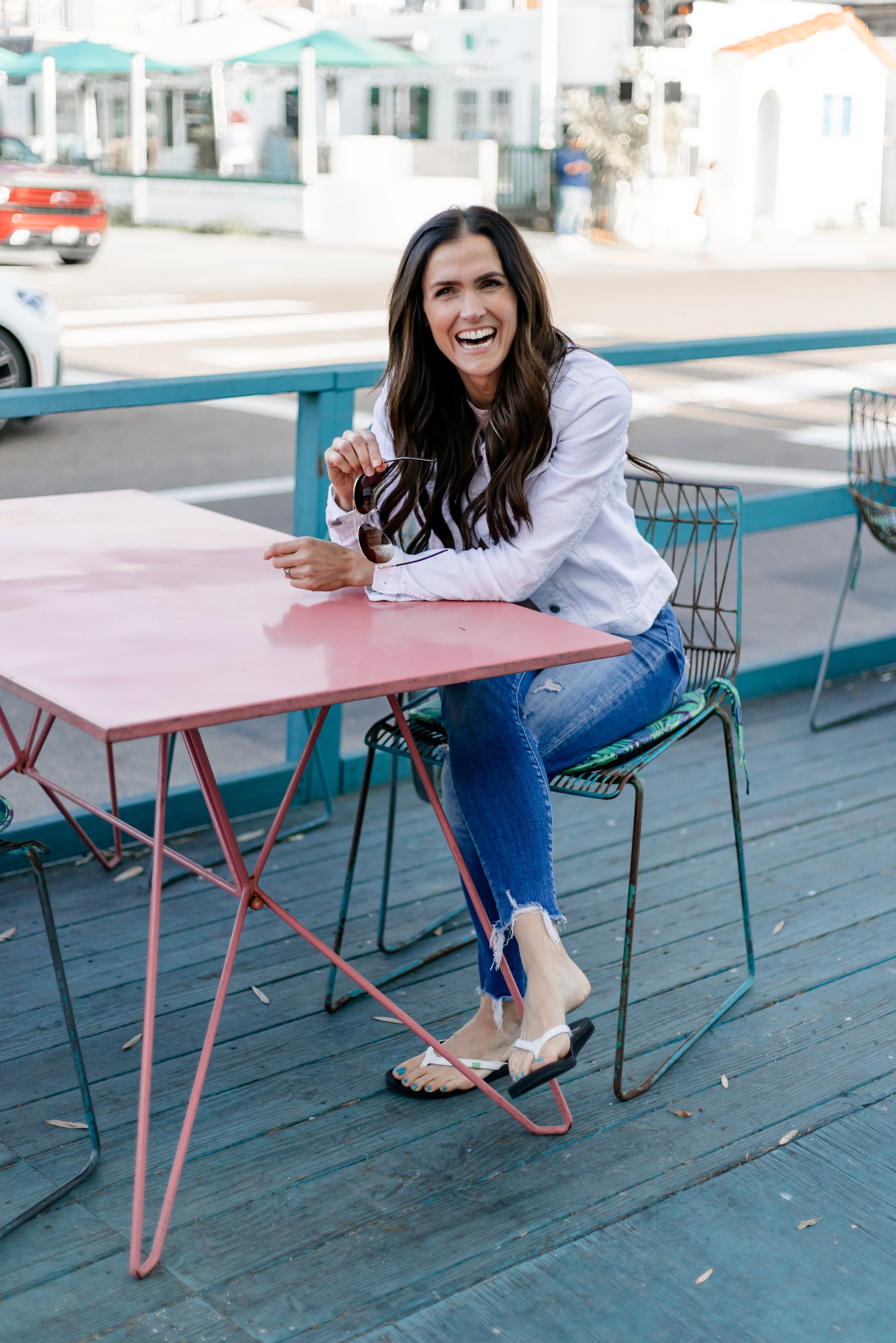 Woman sitting at outdoor cafe table, smiling and laughing, holding sunglasses, wearing a white denim jacket and ripped blue jeans, on a wooden deck with a street and cars in the background.