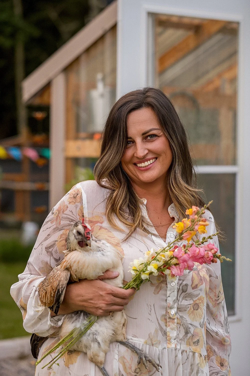 photo of woman holding a chicken and smiling in front of her greenhouse homesteading