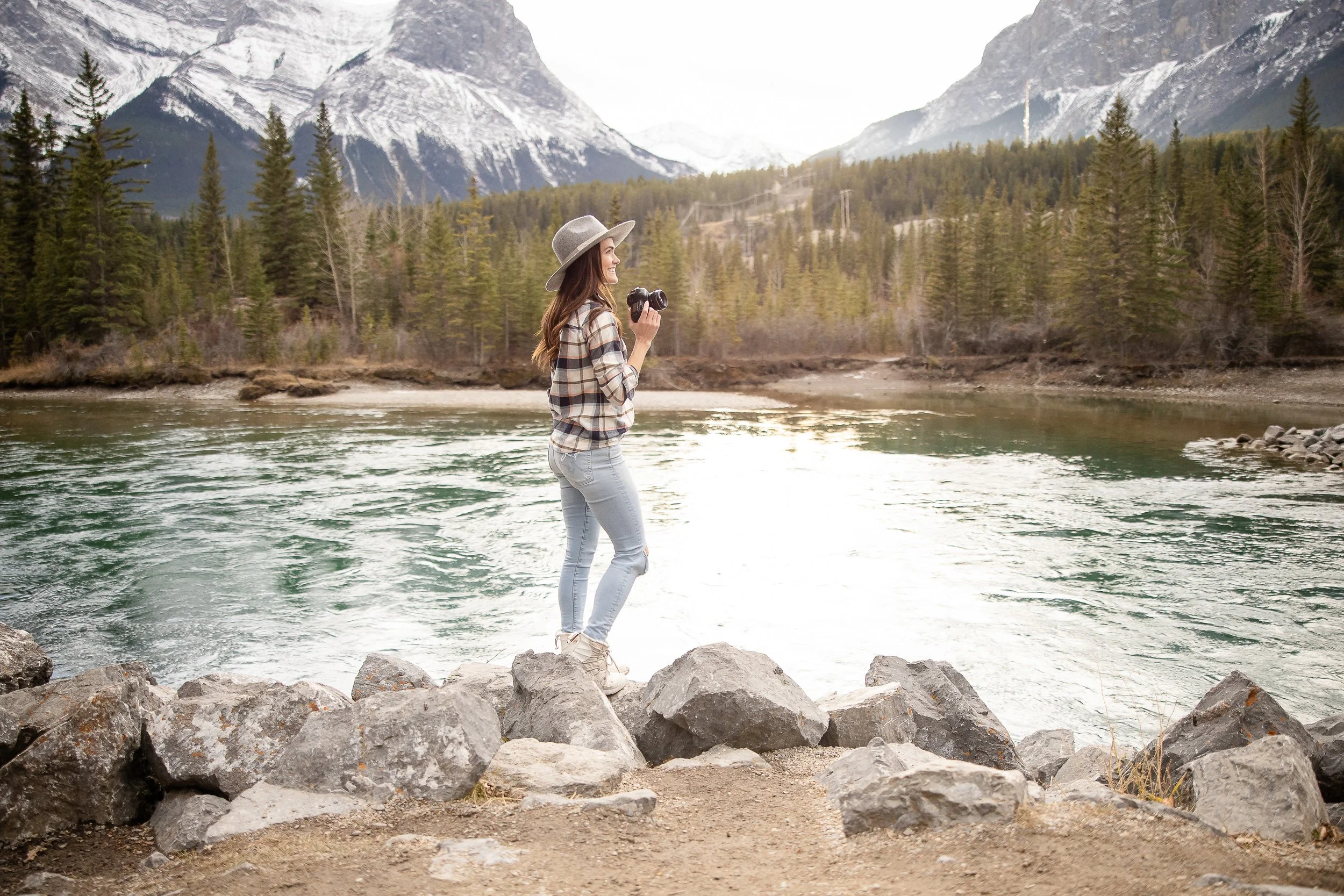A young woman stands on rocks by a river, holding binoculars and looking at the scenic mountain view with snow-capped peaks and trees.