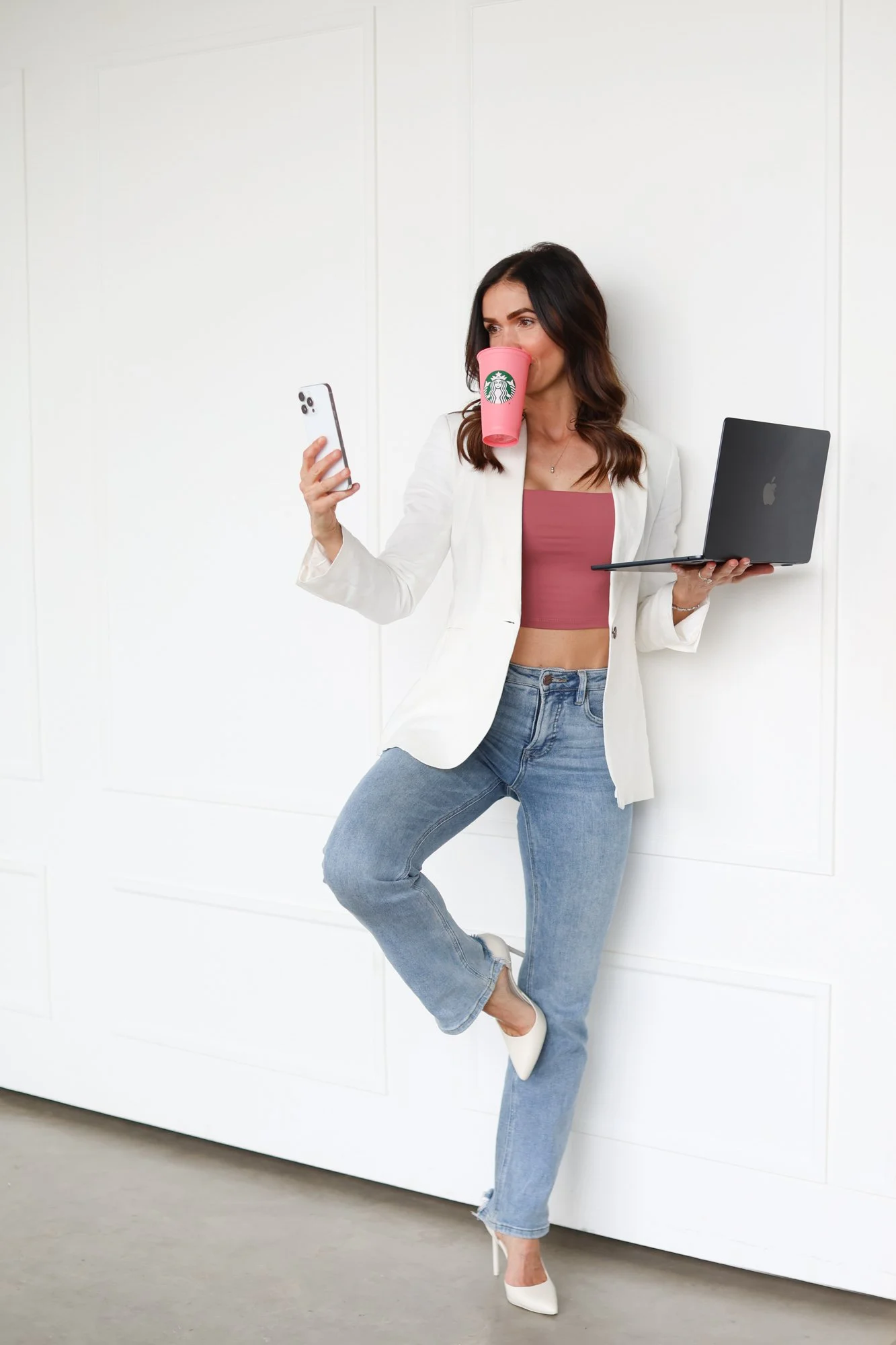 photo of woman with coffee cup in mouth while balancing on one foot holding a phone in one hand and computer in the other, jeans and a pink top