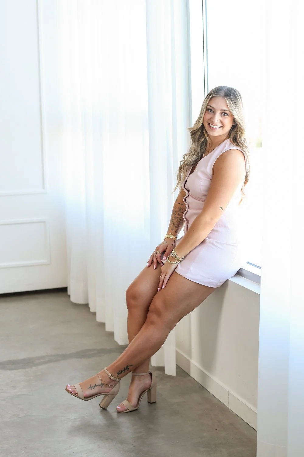 woman leaning against a window with hands on her lap in a light pink button down dress and blonde hair