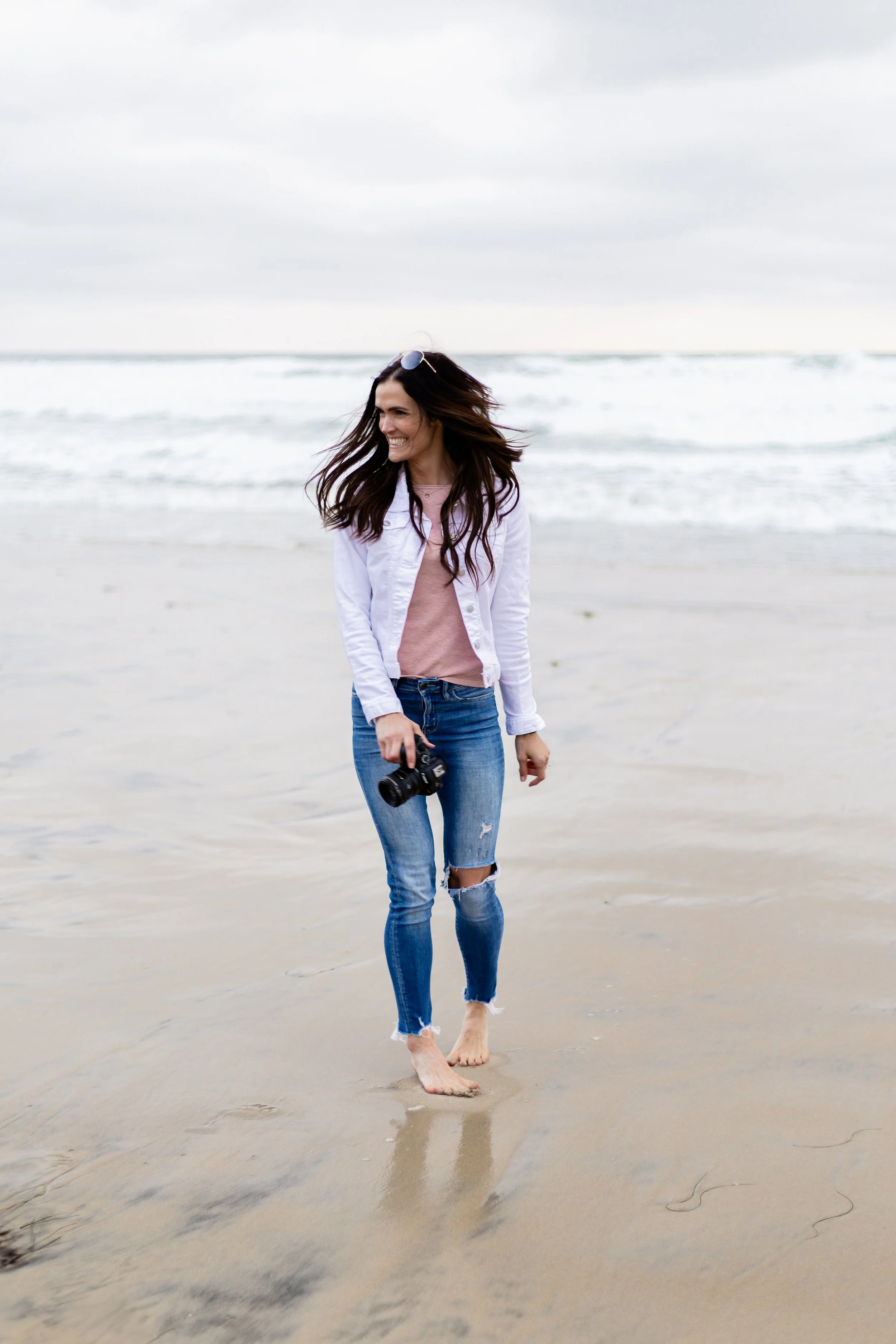 Woman walking barefoot on the beach, holding a camera in her right hand, smiling, with overcast sky and ocean waves in the background.