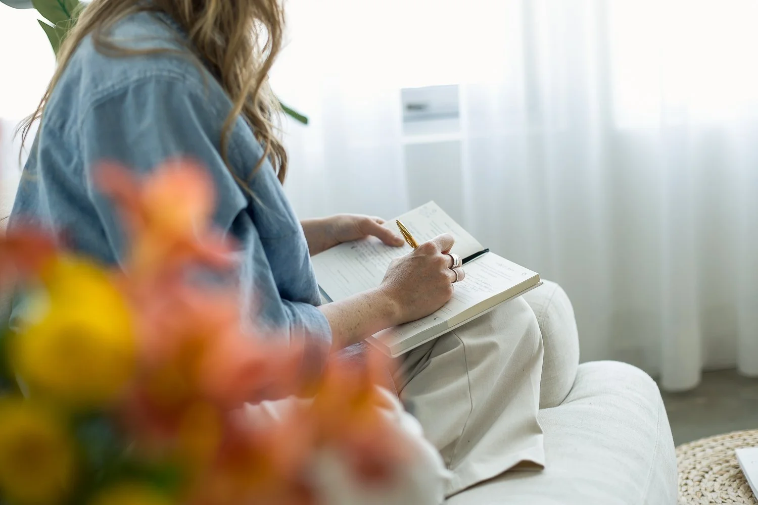 woman sitting in chair journaling with flowers blurred in foreground and white curtains behind her