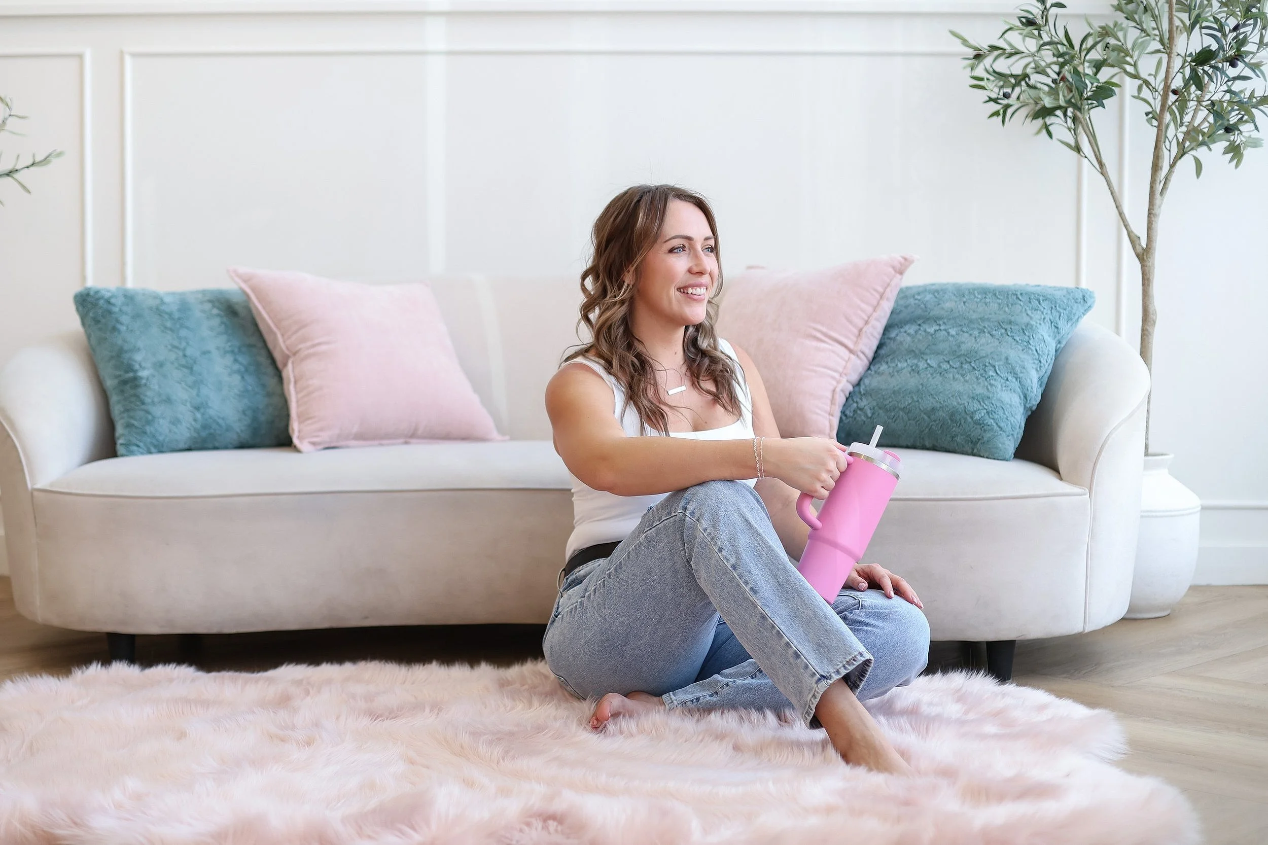 A woman sitting on a pink fluffy rug in front of a white sofa with pink and blue pillows, holding a pink insulated drink container, and smiling in a bright living room with a large plant in the background.